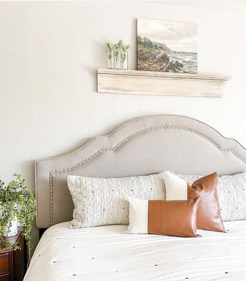 A neatly made bed with Modern Farmhouse bedding and decorative pillows against a headboard, flanked by potted plants, under a shelf displaying a serene landscape painting.