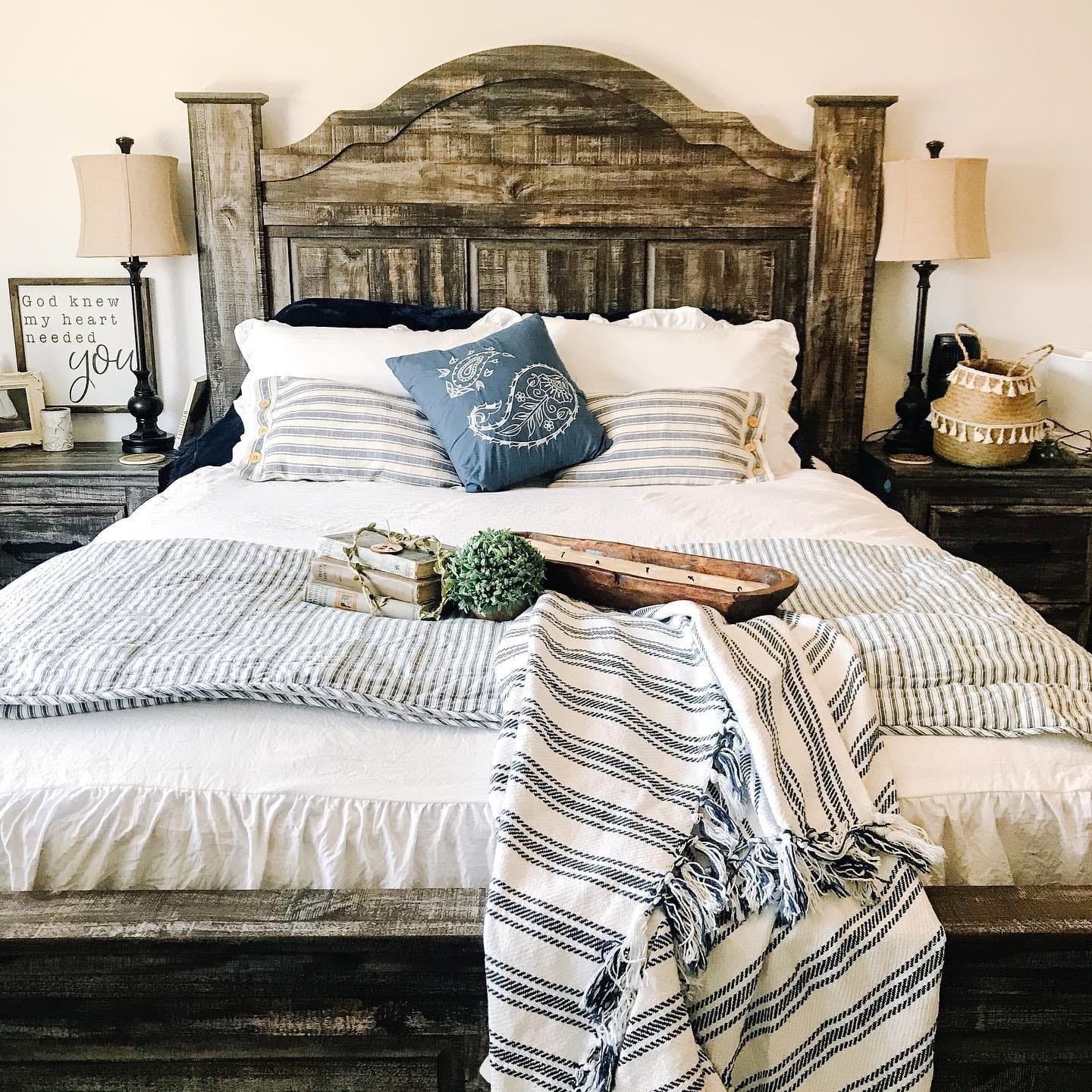 Rustic-style bedroom with wooden headboard, modern farmhouse bedding, two table lamps, and decorative items including a tray and a woven basket.