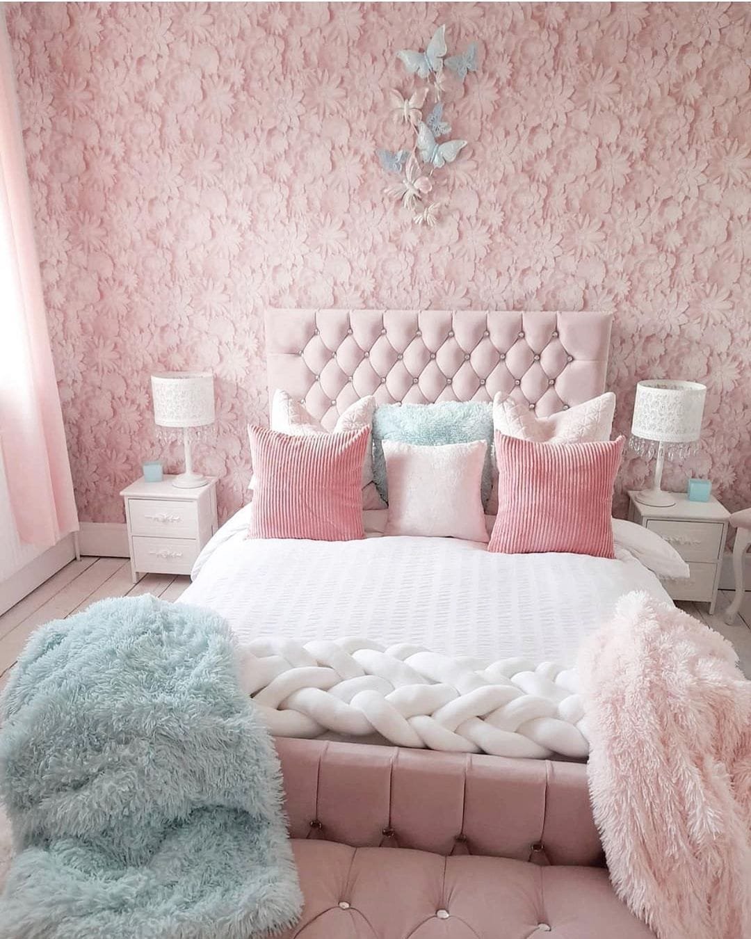 A pink-themed bedroom featuring a tufted bed with matching pillows, fluffy blue and pink blankets, and magical floral wallpaper.