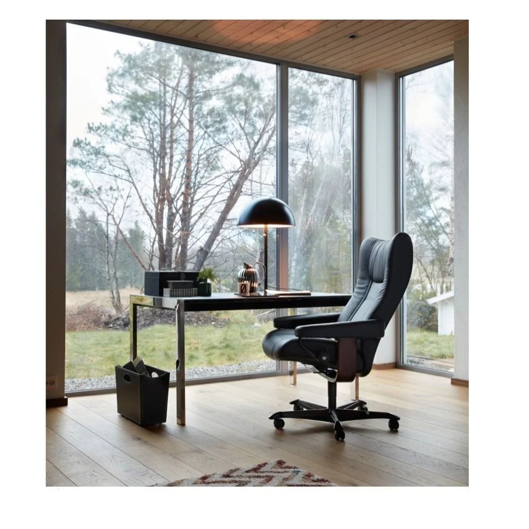 A modern home office with large windows overlooking a forest, featuring a black desk, swivel chair, and a blue lamp on how to optimize your workspace for maximum efficiency.