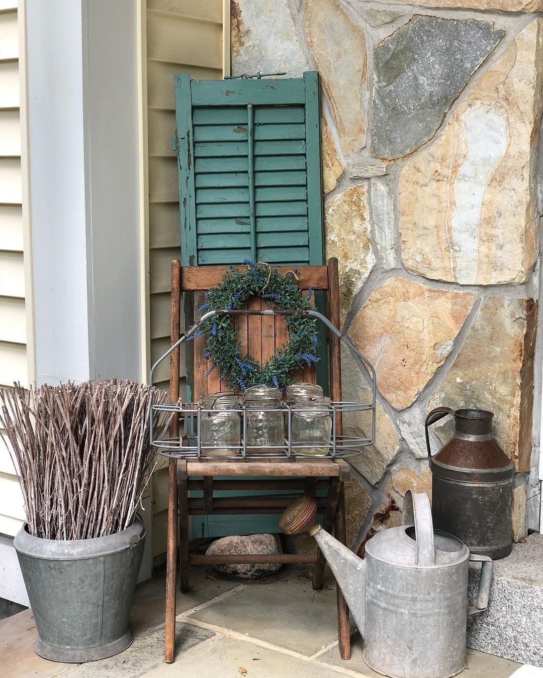 A rustic porch display featuring a wooden chair adorned with a wreath, surrounded by metal buckets, a watering can, and a bundle of twigs creates unique vignettes to decorate an amazing front porch.
