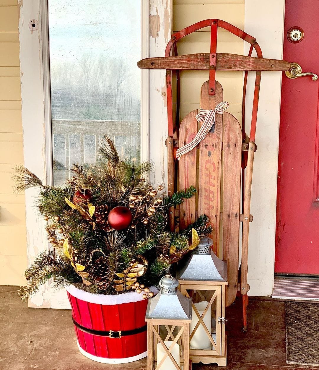 Vintage wooden sled and ski propped against a red door, next to a red bucket with a unique Christmas pine and two white lanterns on an amazing front porch.