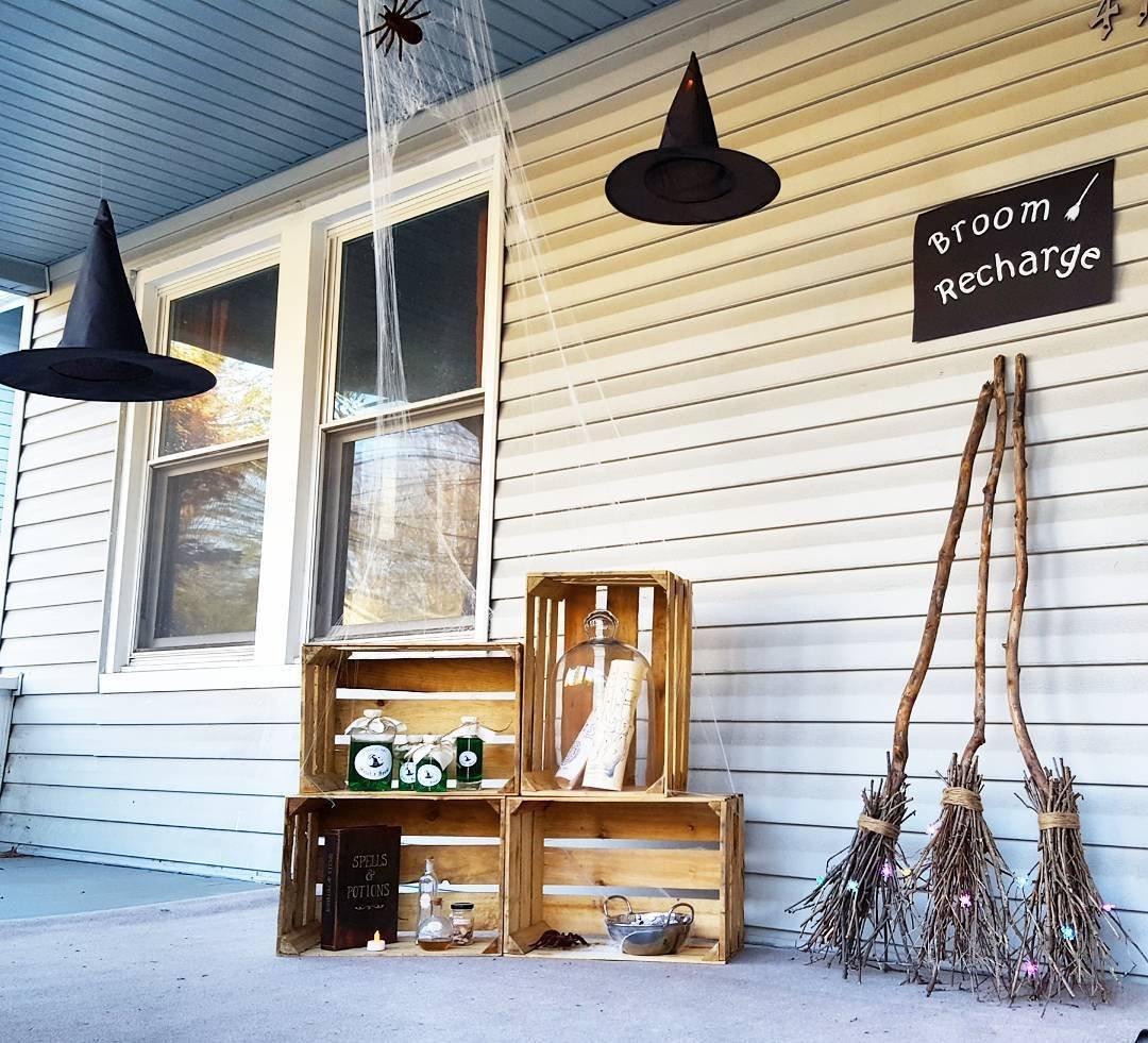 A front porch decorated for Halloween with unique vignettes, a "broom recharge" sign, and witches' hats hanging above crates displaying jars and bottles.