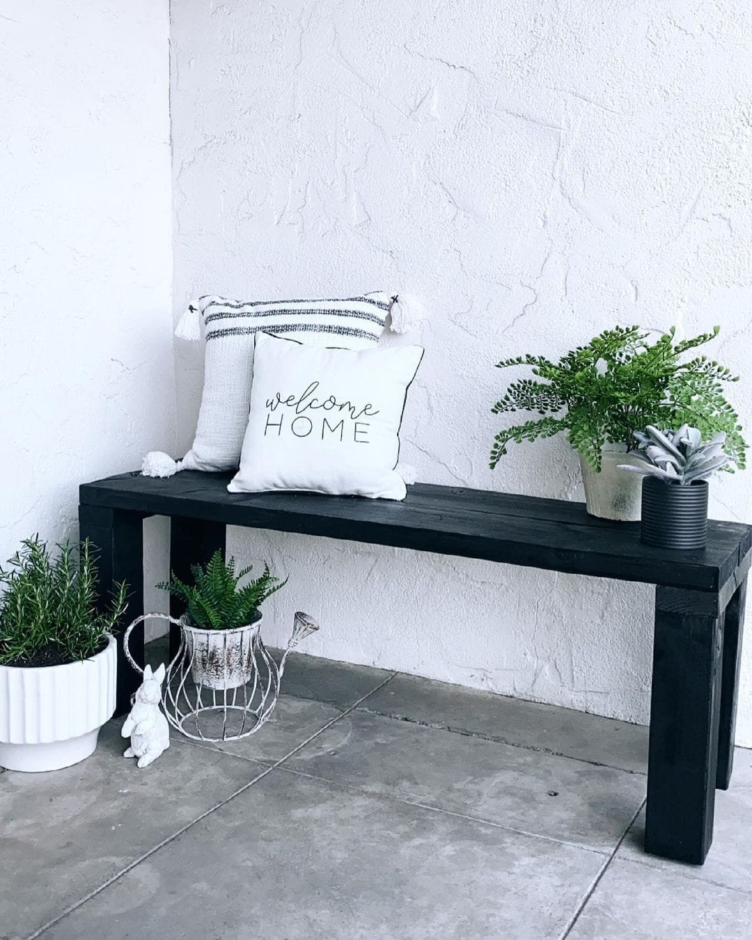 A cozy outdoor bench with a "welcome home" pillow and several potted plants, including ferns, on and beside it, creating unique vignettes to decorate an amazing front porch.