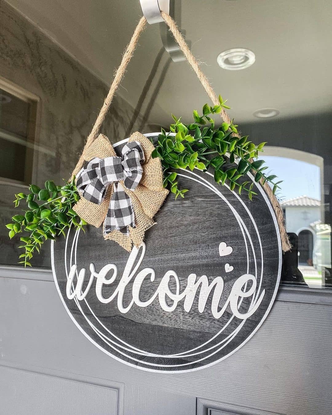 A circular "welcome" sign hanging on a glass door, decorated with green foliage and a burlap bow with a gingham pattern, creates unique vignettes to decorate an amazing front porch.