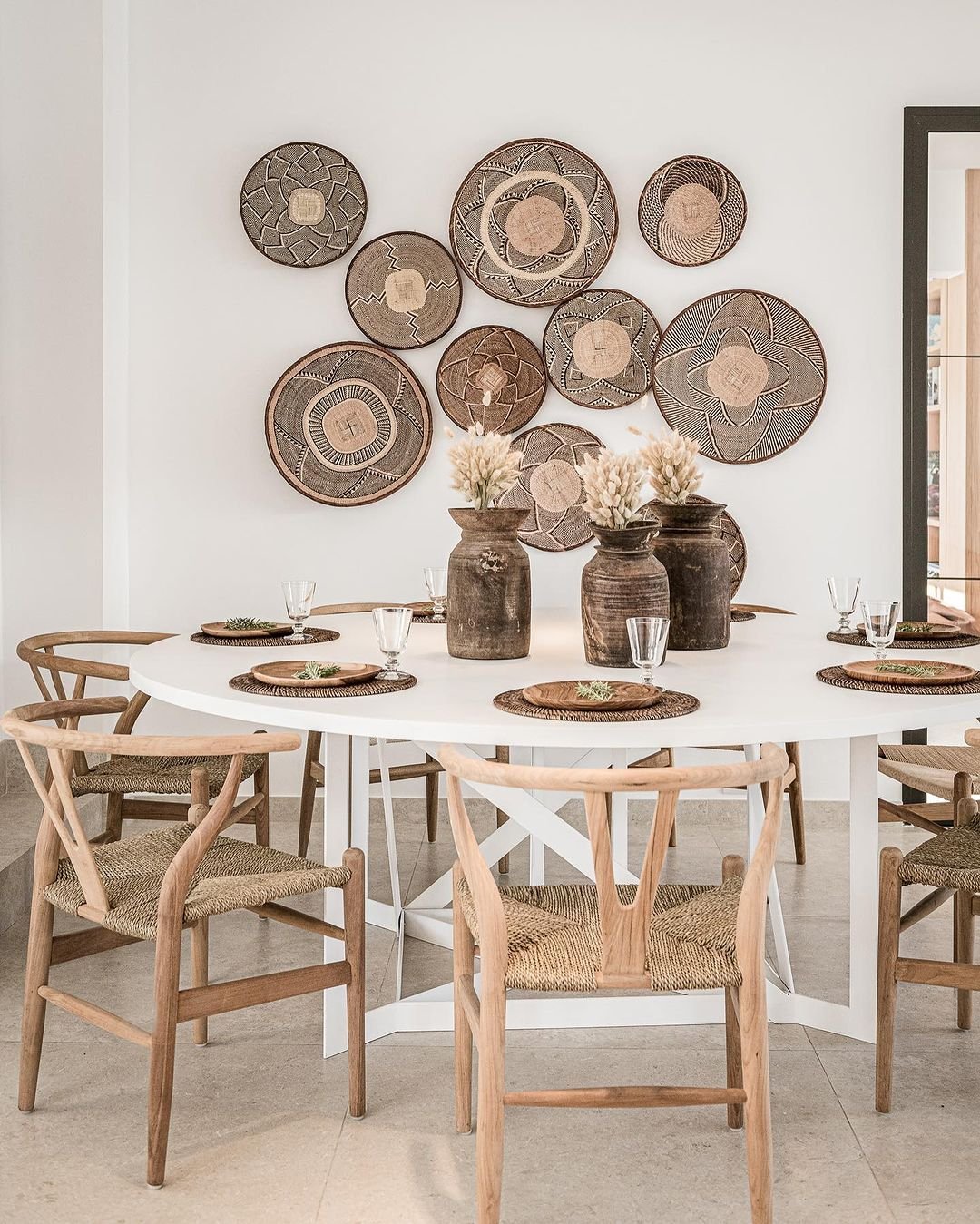 A dining area with a round white table, six wooden chairs, woven placemats, glassware, and three large vases containing dried flowers. Bohemian wall basket decor in traditional to contemporary styles is displayed on the white wall behind.
