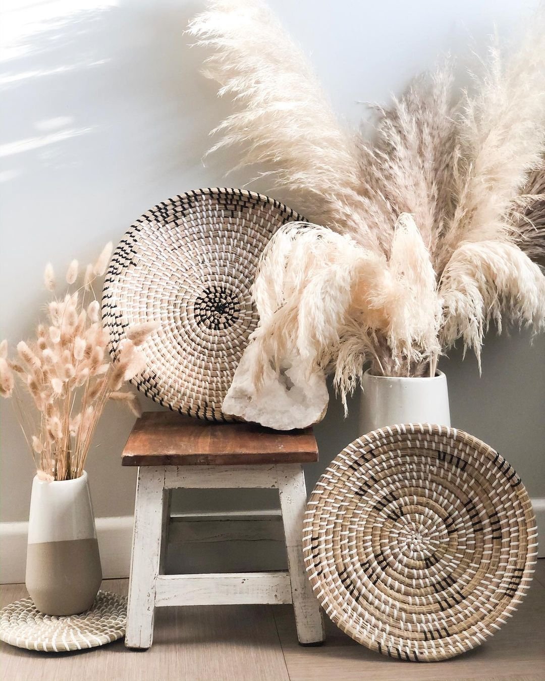 A small wooden stool is decorated with woven baskets and vases holding dried pampas grass and beige grass, creating a charming bohemian wall basket decor against a light-colored wall.
