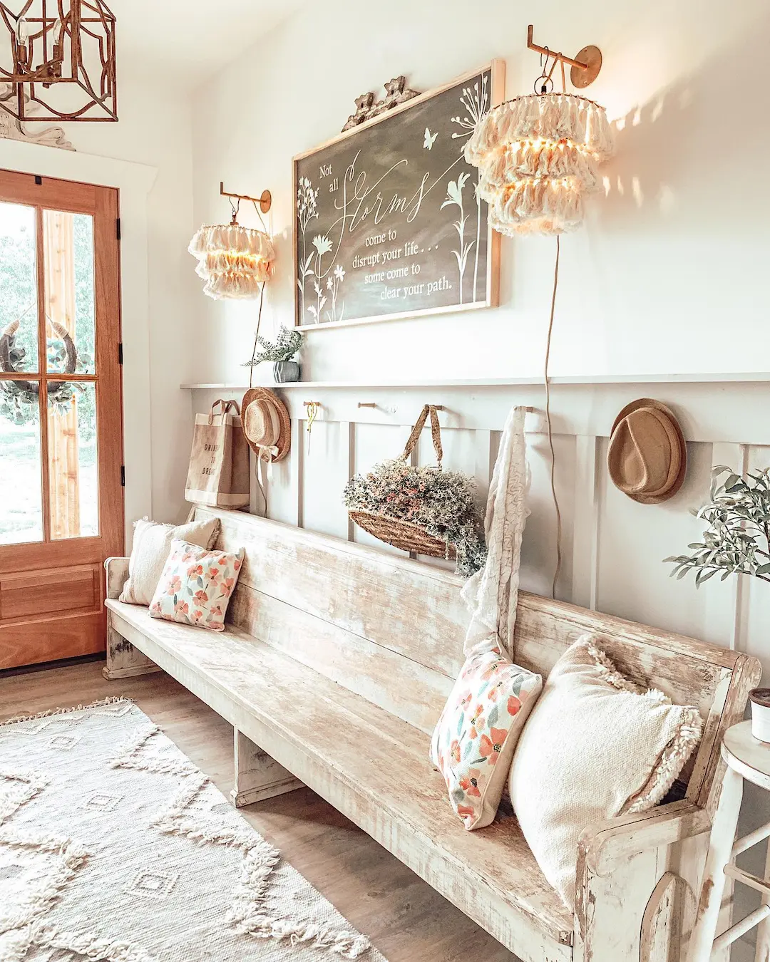 A rustic entryway shows how to style your entryway with a farmhouse bench, featuring a distressed wooden seat, patterned pillows, hats on hooks, a hanging wreath, and soft lighting from two wall lamps beneath a decorative sign.