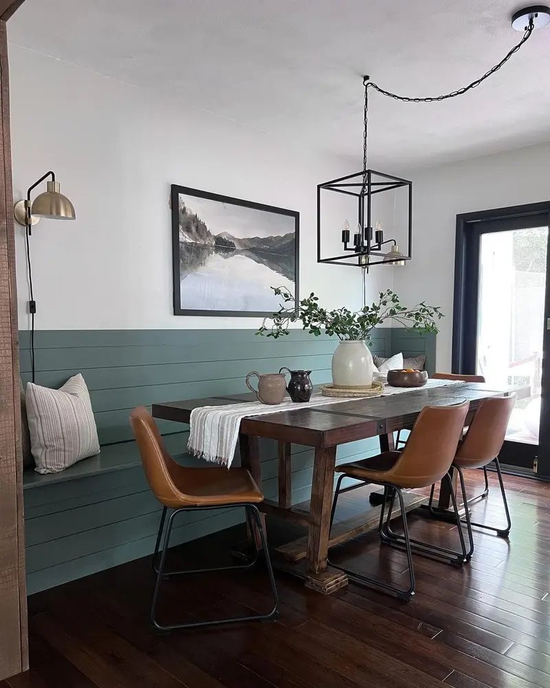 A modern dining room featuring a rustic wooden table, banquette seating, a wall-mounted landscape painting, and contemporary lighting fixtures.