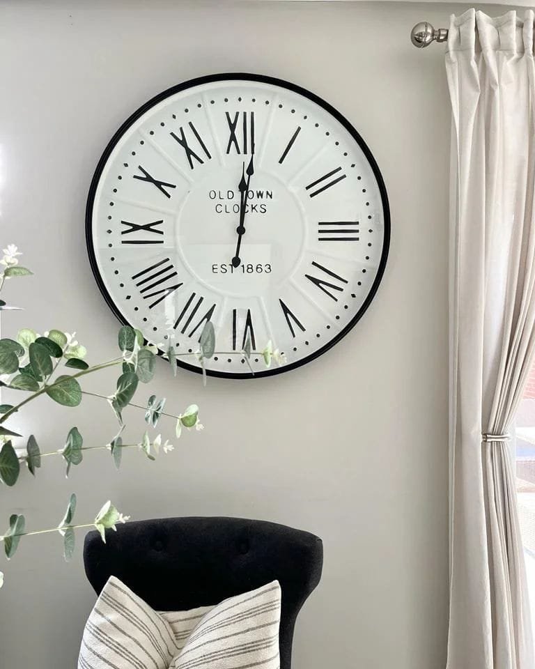 Large wall clock with Roman numerals, labeled "Old Town Clocks, Est 1863", on a light gray wall beside a window with white curtains and a potted plant for a funky decor.
