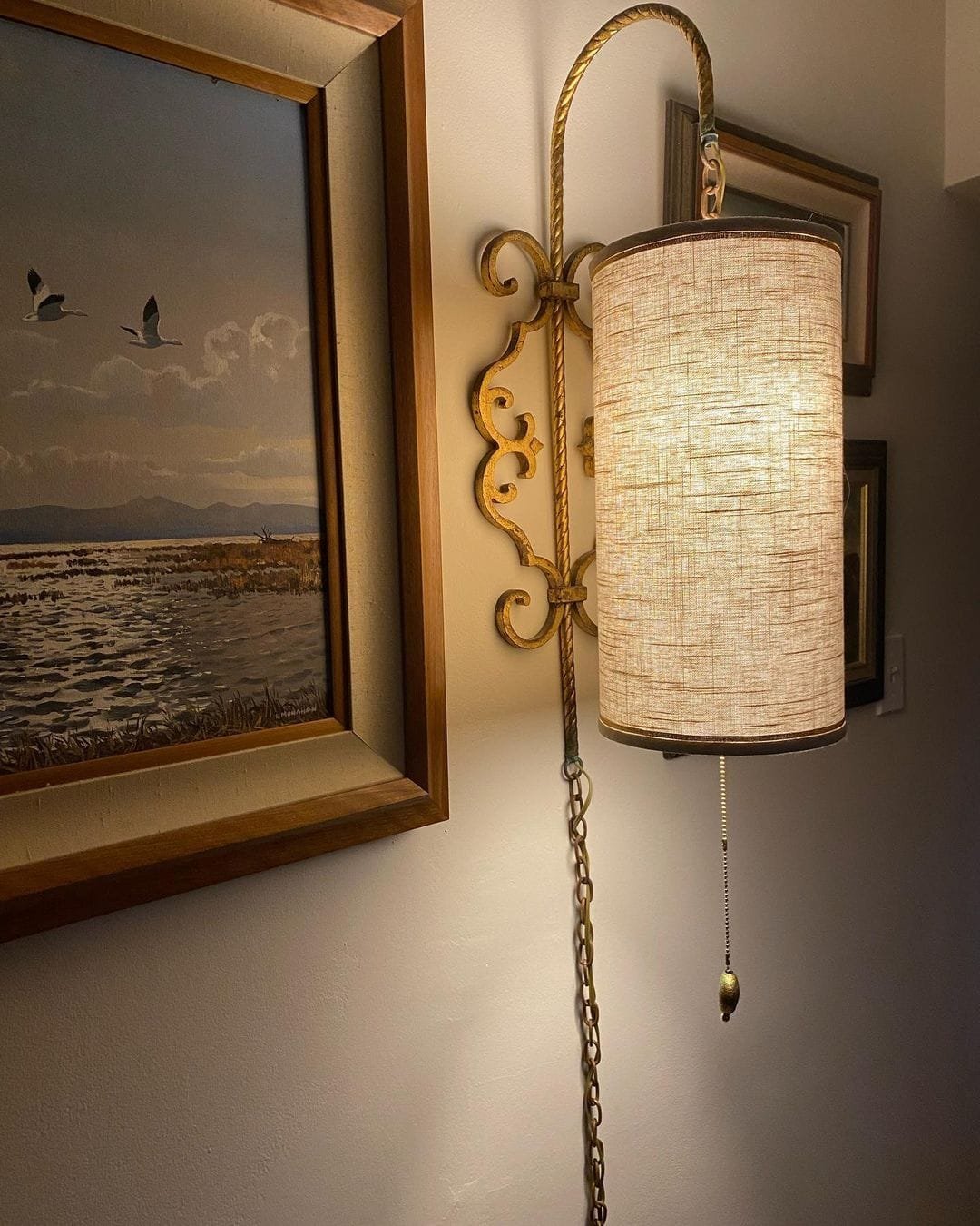 A wall-mounted light fixture with a beige shade and a pull chain, next to a framed painting of a seascape with birds in a vintage frame.