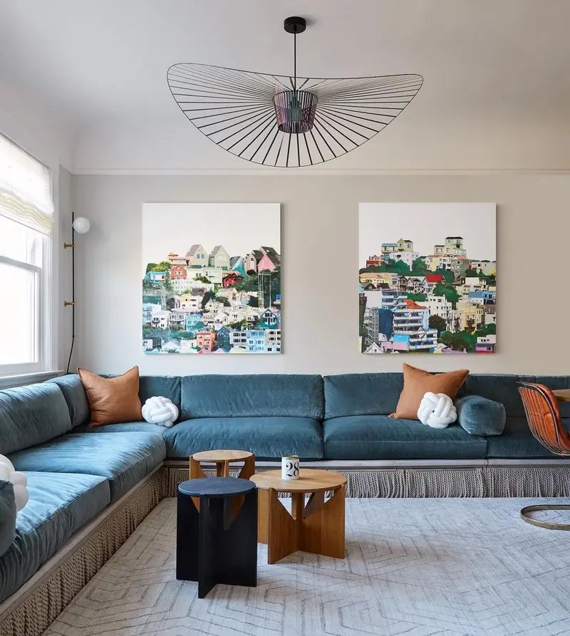A modern living room with a large blue sectional sofa, two vibrant town-themed paintings on the wall, a unique ceiling light fixture, and wooden banquette seating.