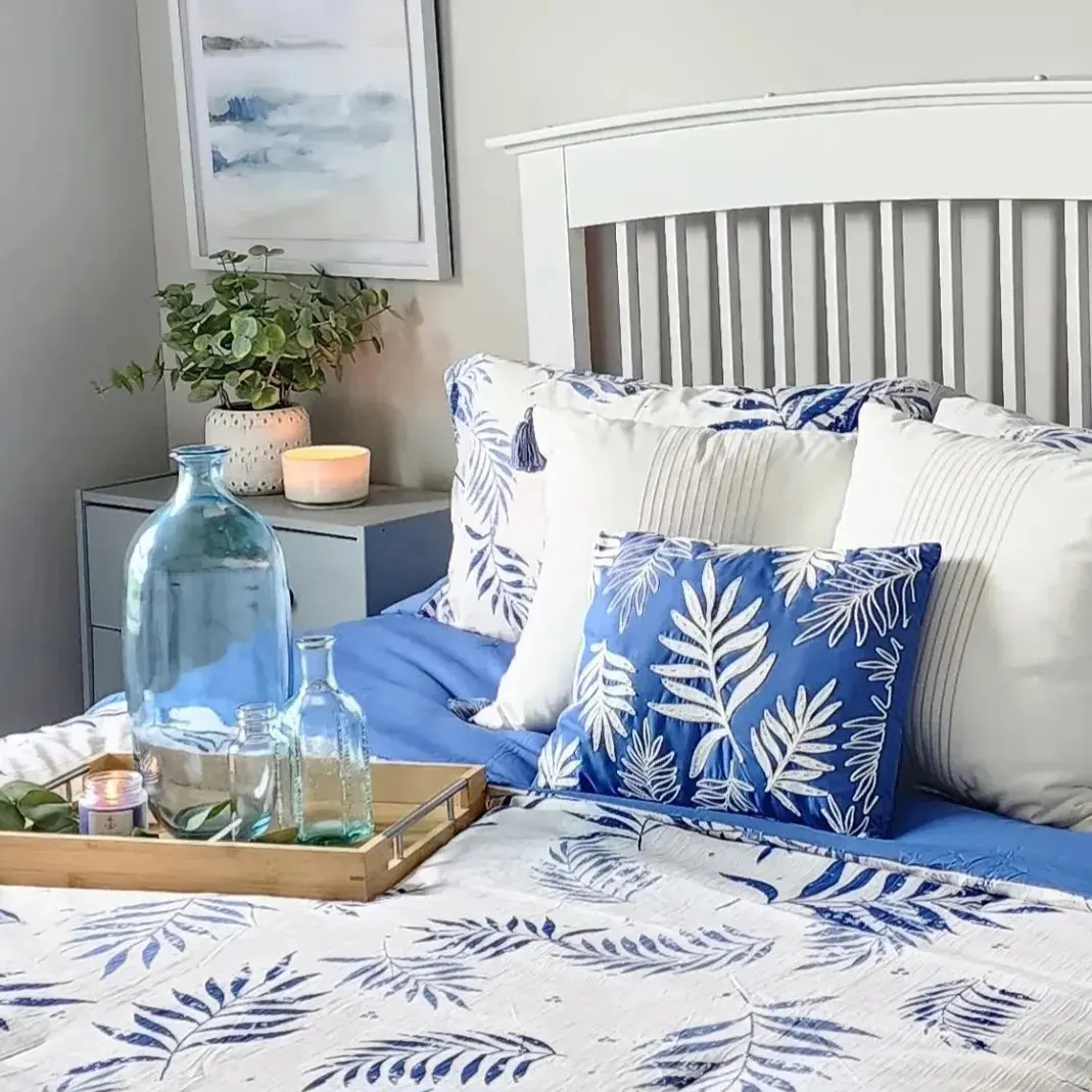 A neatly made bed with blue and white leaf-patterned bedding, a wooden tray with glass bottles and a candle, plus a plant on the nightstand—showcasing summer colors: how to incorporate bright hues into your home decor.