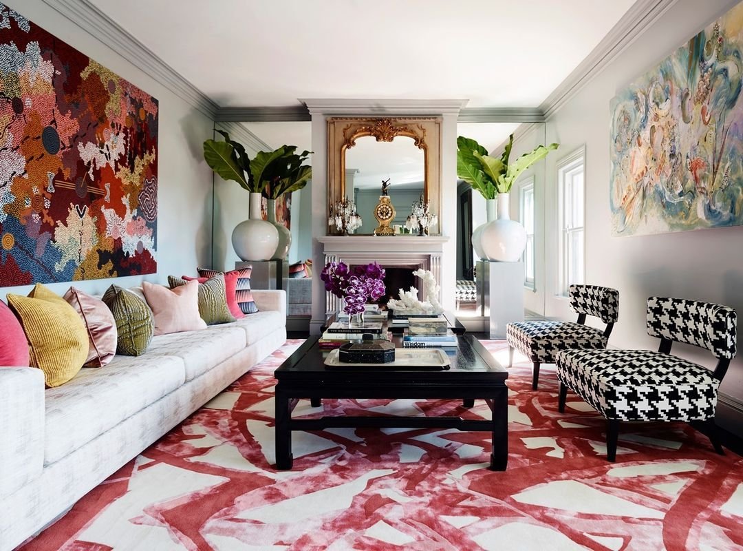 A living room with colorful abstract wall art, large leafy plants, a mirror above a fireplace, and a white sofa with multicolored cushions exemplifies maximalist decor: how to balance eclectic and overwhelming. Patterned chairs and a black coffee table on a red and white rug complete the vibrant space.