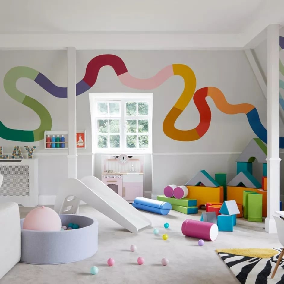 A colorful, vibrant children's playroom designed to be both playful and practical, with a mural of wavy lines, a slide, various play blocks, and toys scattered around the room.
