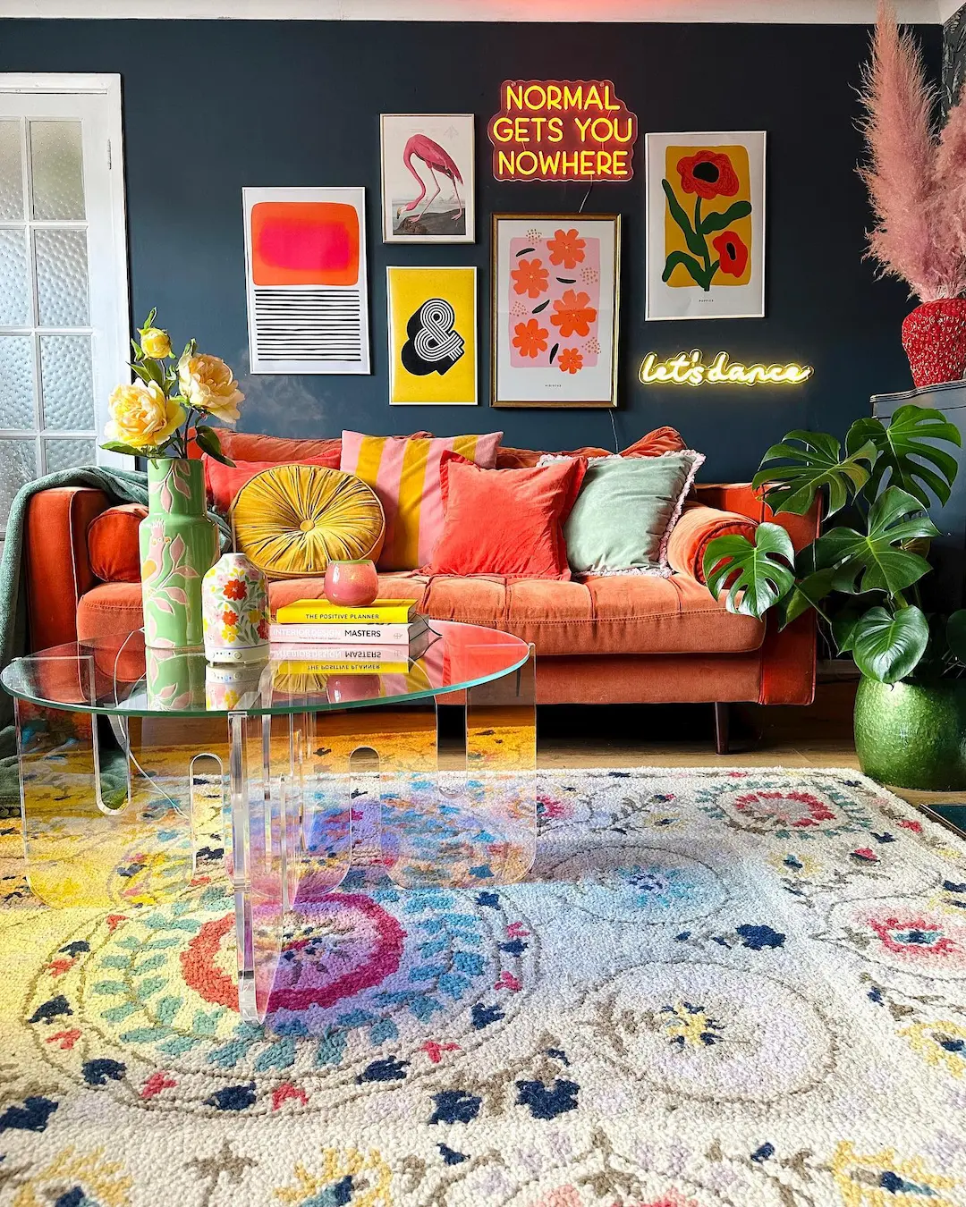 A colorful living room with a bright orange sofa, vibrant cushions, wall art, neon signs, glass coffee table, and houseplants showcases maximalist decor—perfectly balancing eclectic style without becoming overwhelming.