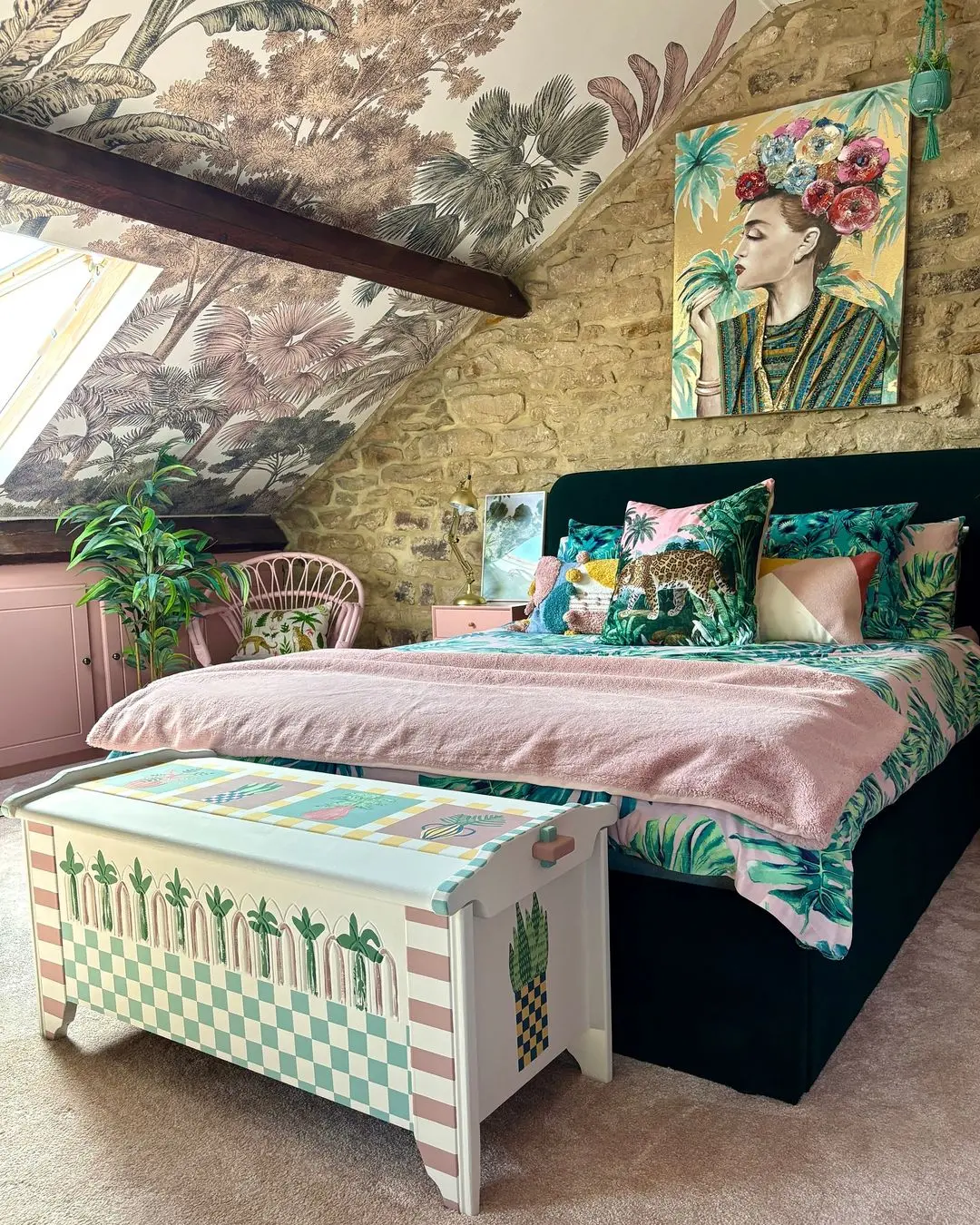 A bedroom with a sloped ceiling, stone wall, and botanical wallpaper showcases maximalist decor: how to balance eclectic and overwhelming with green and pink accents, decorative pillows, and a painted storage chest at the foot of the bed.