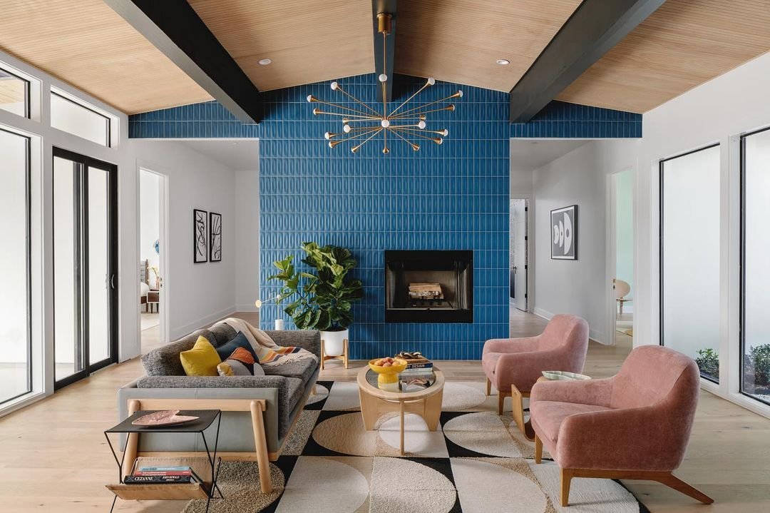 Minimalist living room with blue tiled fireplace, pink armchairs, wooden ceiling, and large glass doors.