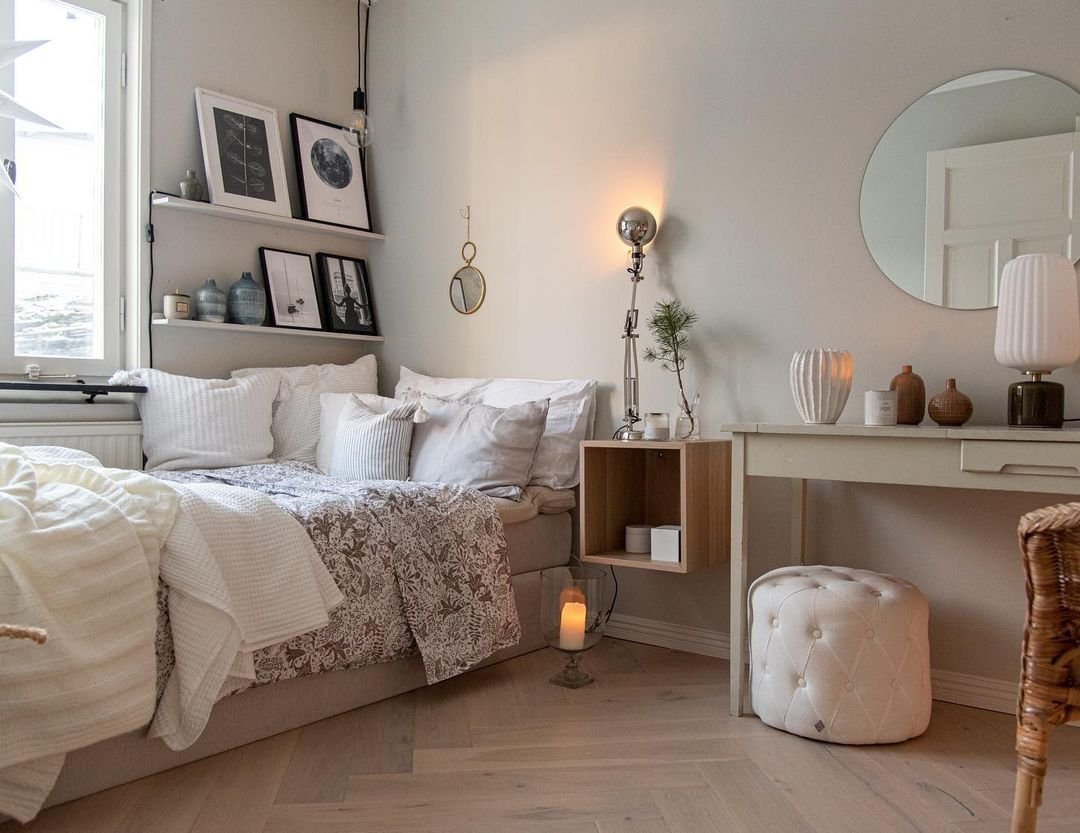 A cozy Scandinavian bedroom with a neatly made bed, beige walls, a round mirror, and decorative items on a shelf and walls.