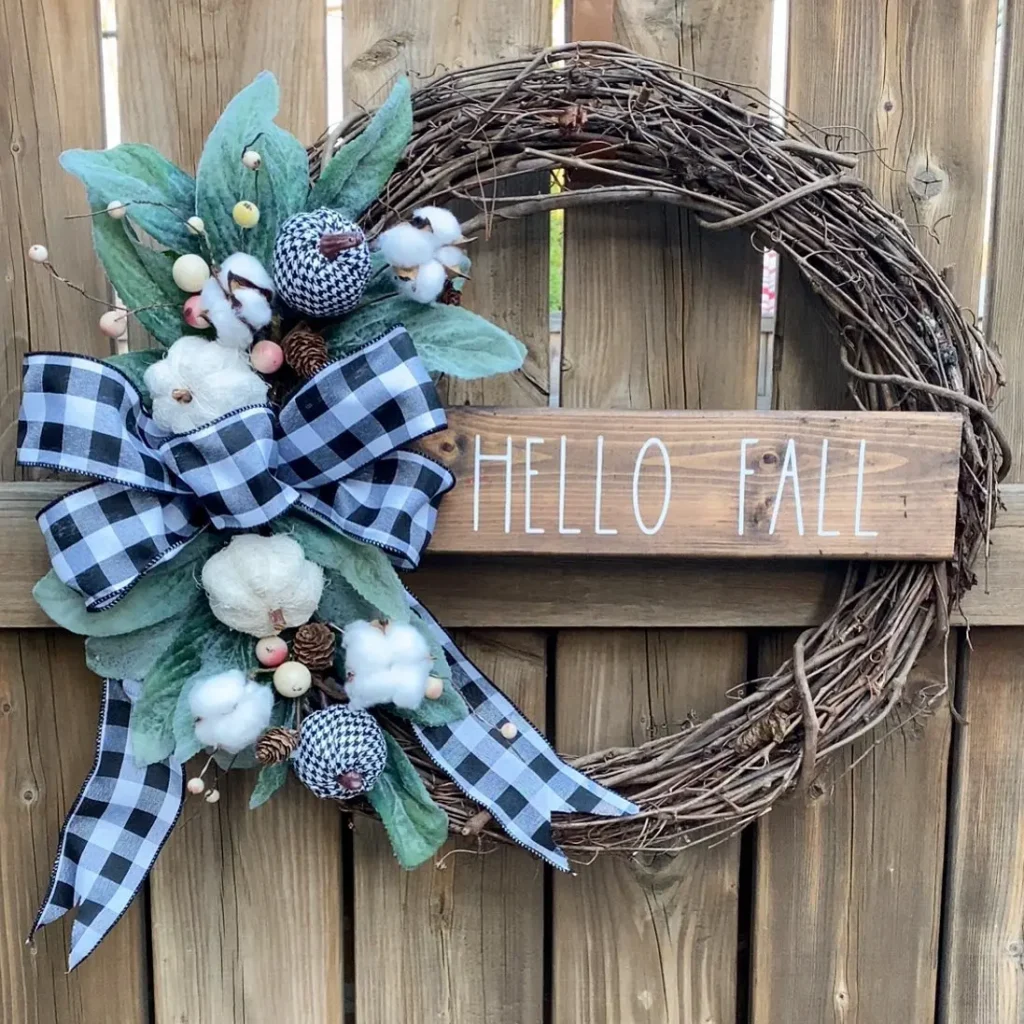 A grapevine wreath decorated with cotton, leaves, and a black-and-white plaid bow shows how to embrace buffalo plaid in fall decor, featuring a "HELLO FALL" wooden sign hanging on a wooden fence.