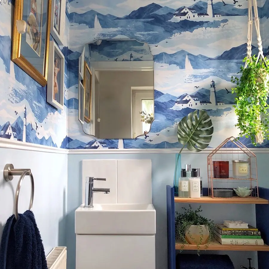 A bathroom with blue and white mountain-themed wallpaper, a white sink, a mirror, a towel on a silver rack, and a shelf holding toiletries and plants creates the perfect coastal bathroom decor for a relaxing oasis.