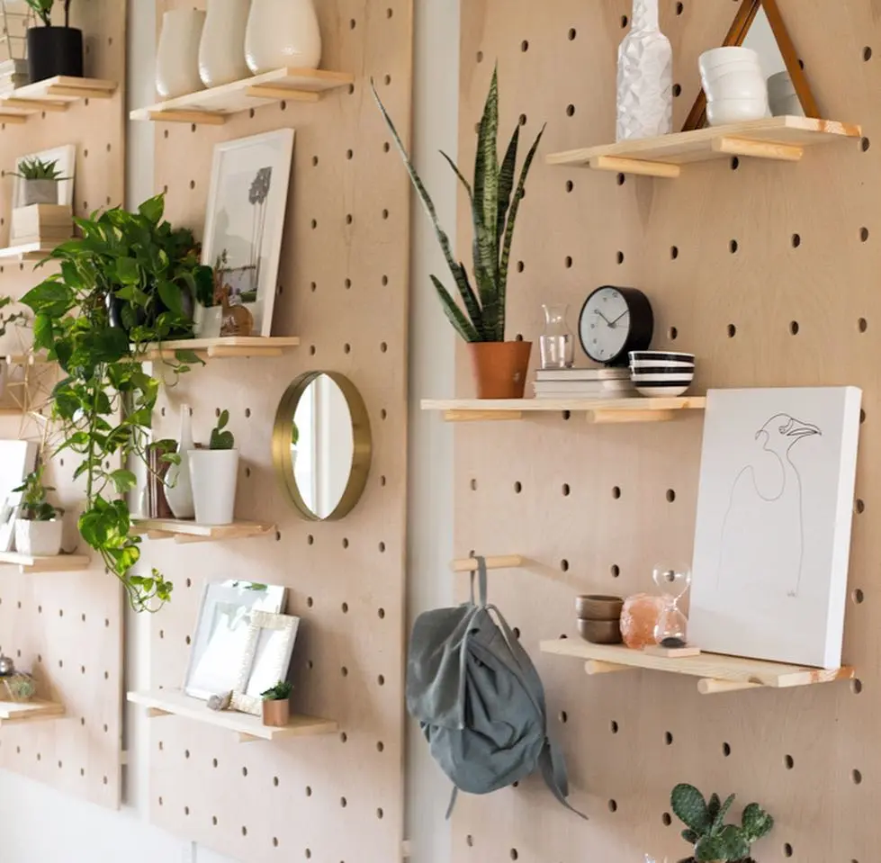 Organization Made Stylish: How to Decorate with a Wall Pegboard—wooden pegboard wall with shelves displaying potted plants, framed photos, art, decor items, a round mirror, and a hanging bag for a chic touch.