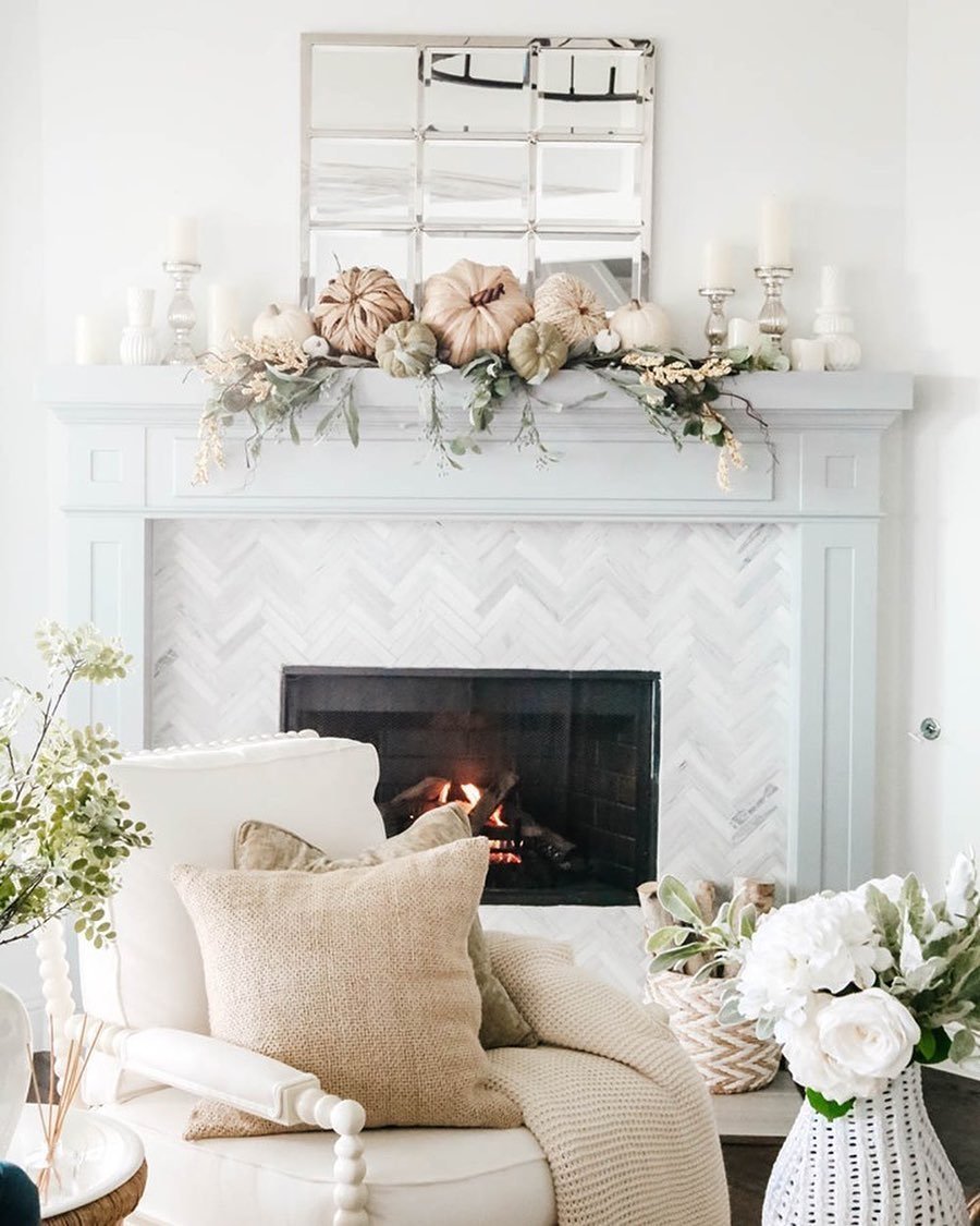 A cozy living room features a lit fireplace with a fall-themed mantel, showcasing neutral fall mantel decor ideas for a chic and sophisticated look, adorned with pumpkins and greenery. A white chair with a beige pillow sits nearby, surrounded by plants and flowers.