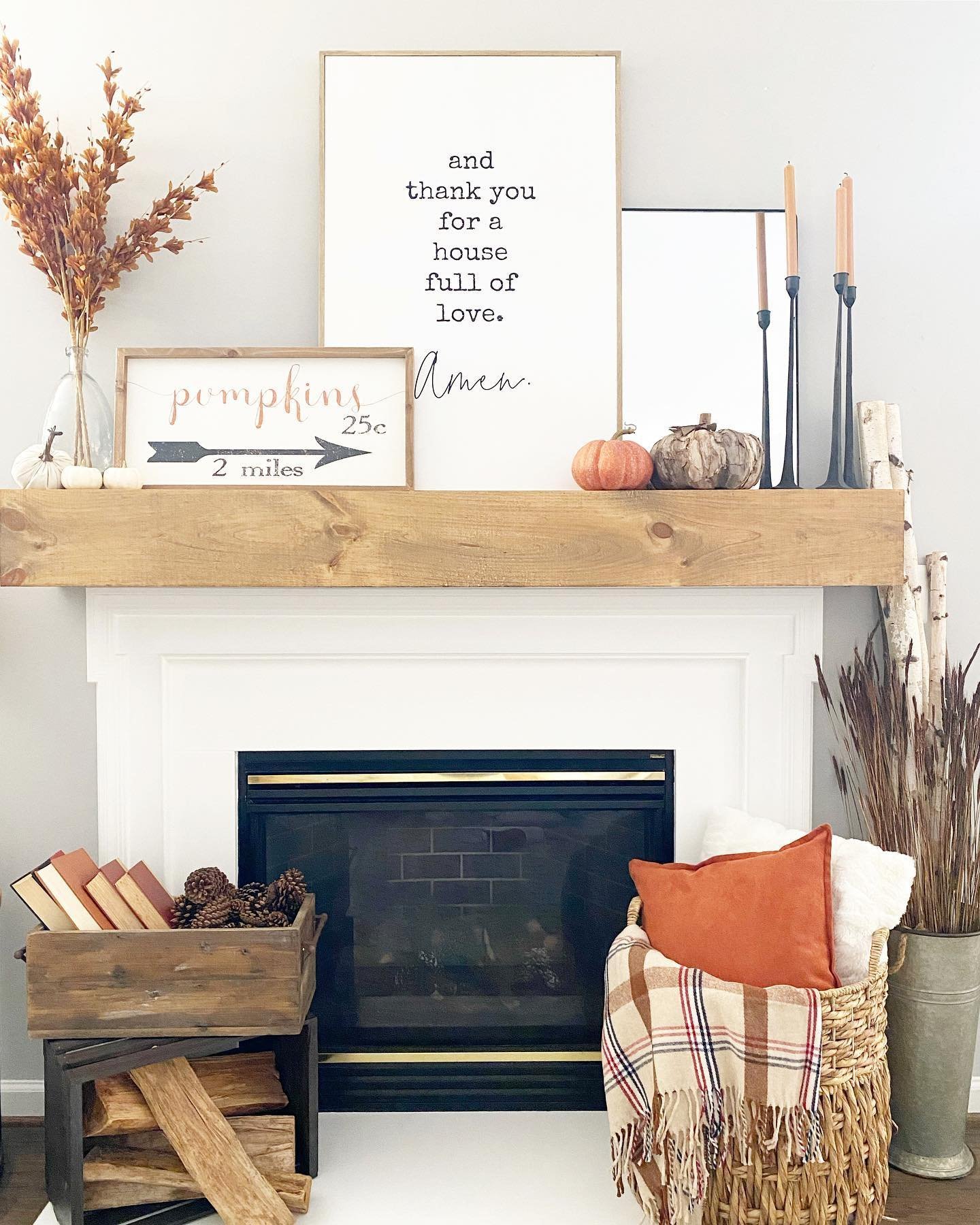 A white fireplace is decorated with framed quotes, candles, pumpkins, and dried leaves—perfect neutral Fall mantel decor ideas for a chic and sophisticated look. Nearby, a basket with an orange pillow and plaid blanket sits next to a wooden crate filled with firewood.
