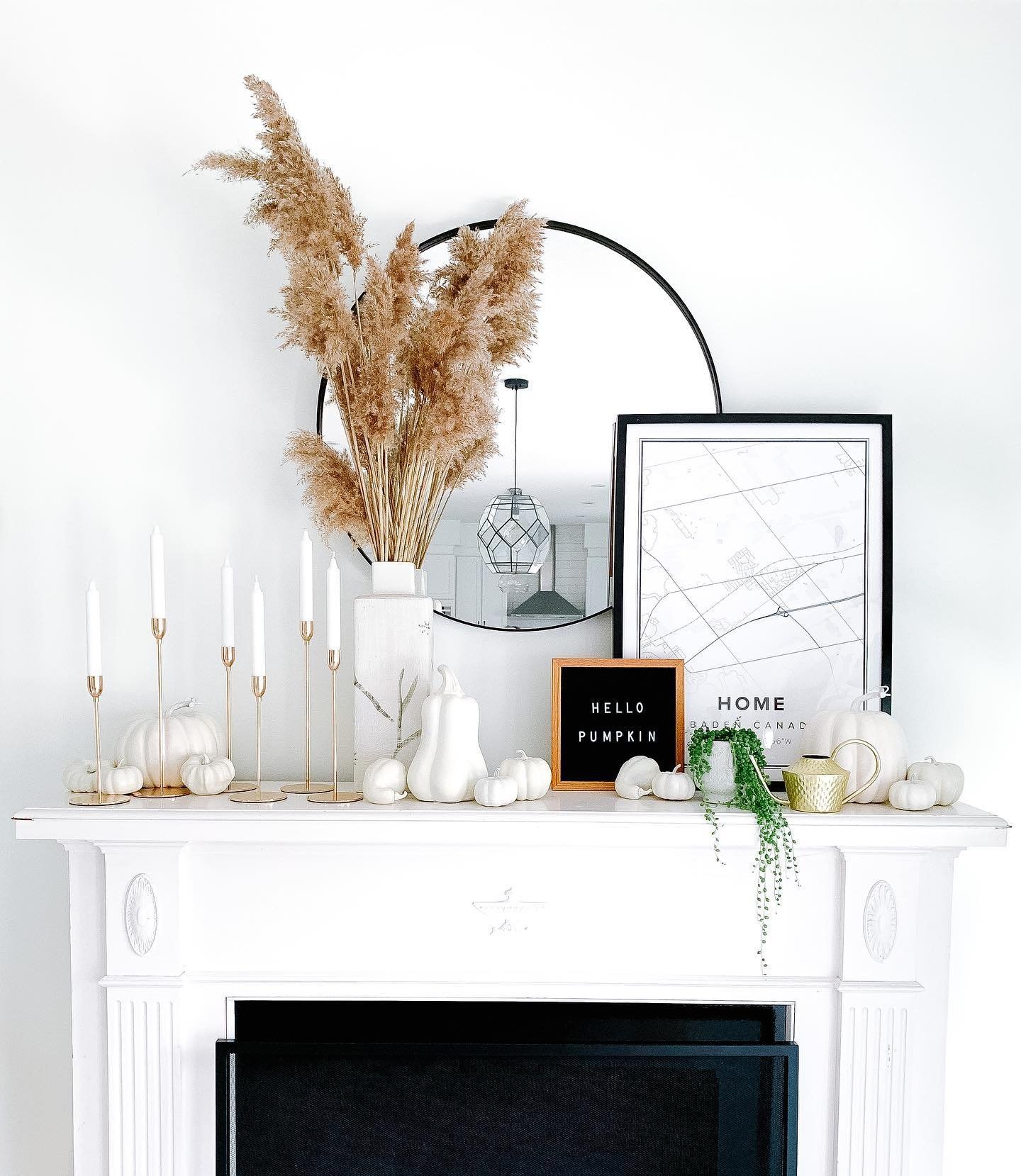 A white mantle is decorated with candles, white pumpkins, framed pictures, a vase of pampas grass, and a letter board reading "HELLO PUMPKIN." A round mirror is mounted on the wall above—a perfect example of neutral fall mantel decor ideas for a chic and sophisticated look.