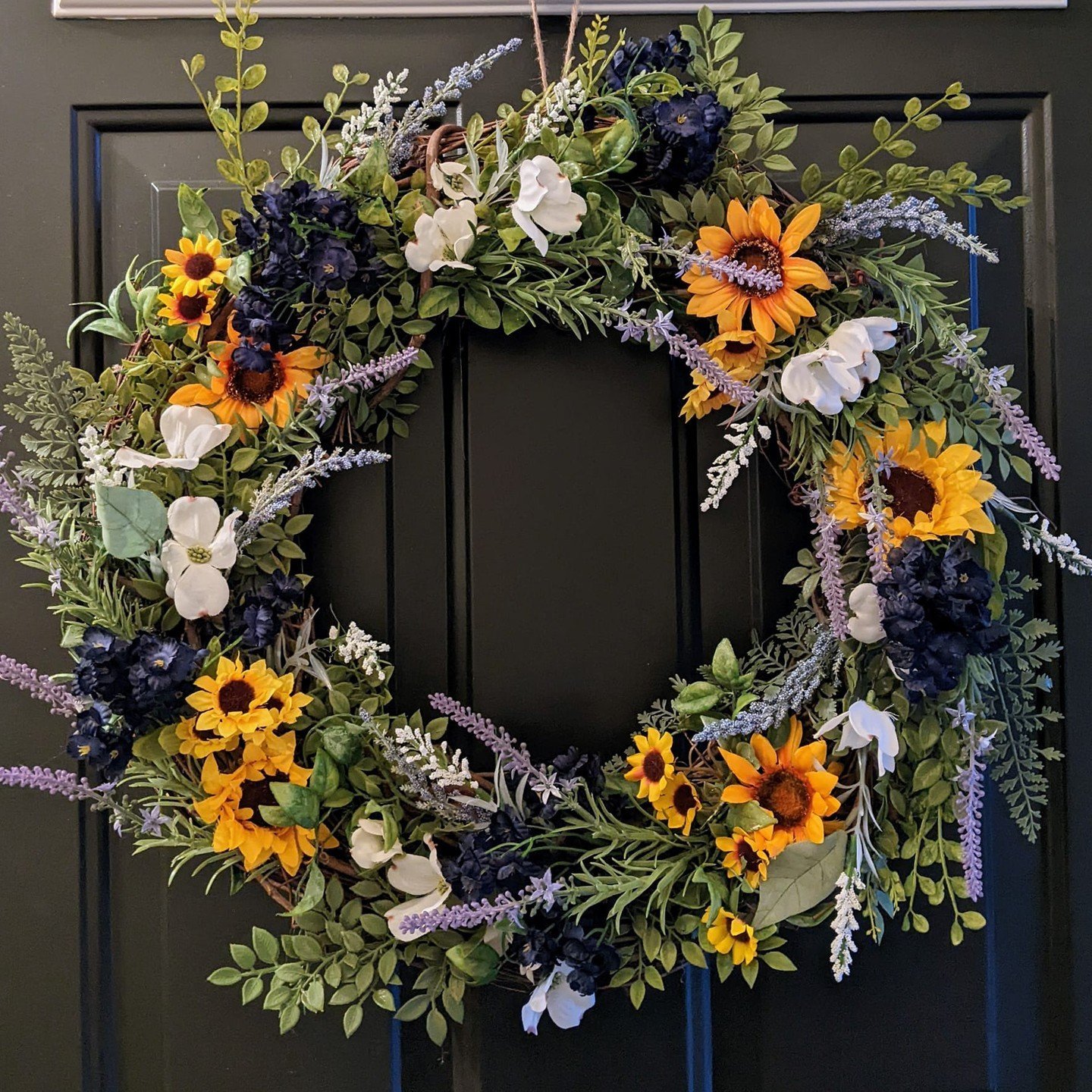 A decorative wreath featuring a mix of yellow sunflowers, white magnolias, and purple flowers surrounded by green foliage is hanging on a dark-colored door. Learn how to make your own fall wreaths with natural materials and scents for a personalized touch.