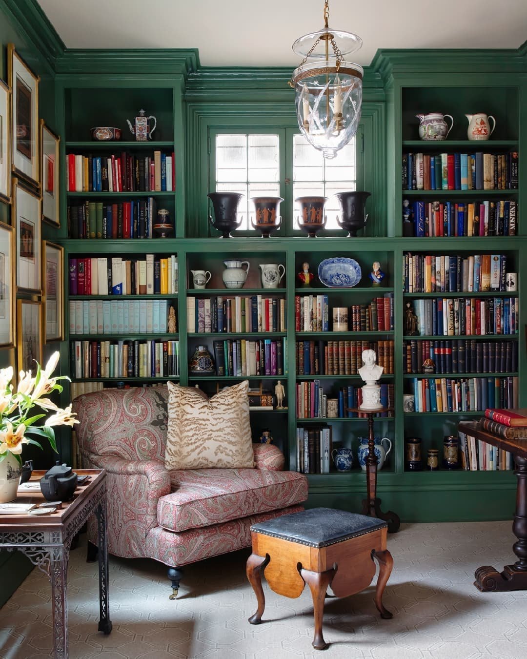 A cozy library with green built-in bookshelves, a patterned armchair, an ottoman, a side table with flowers, framed artwork, and various decorative items on the shelves. A chandelier hangs from the ceiling. Perfect for those seeking ideas from 10 Inspiring Home Office Library Designs for Every Decor Style.