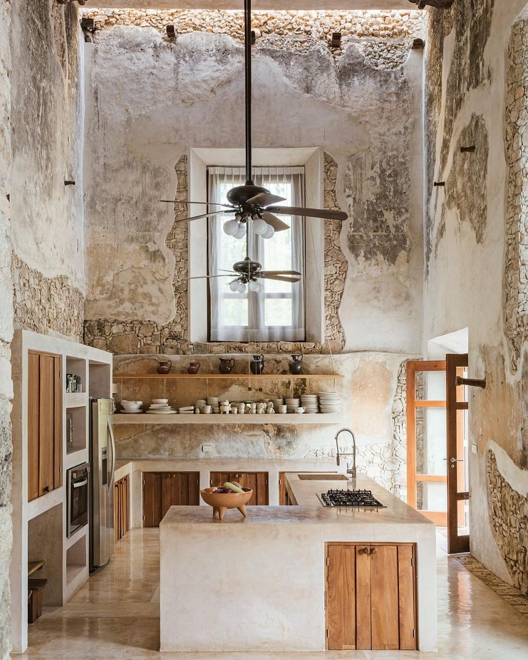 A rustic kitchen with weathered walls, a high ceiling, wooden cabinets, an island with a stove, a ceiling fan, and open shelves displaying dishware. Japandi-inspired design ideas blend minimalism and functionality seamlessly. A door leads outside for easy access to fresh ingredients.