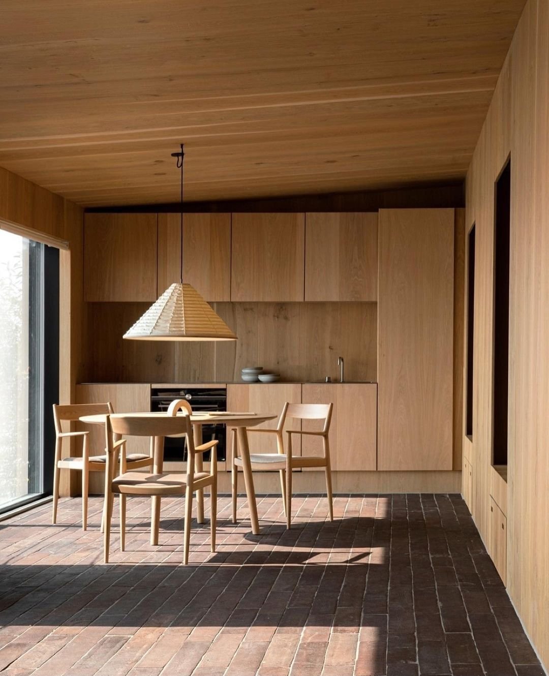 Modern kitchen and dining area with light wood cabinetry, a brick floor, a dining table with four chairs, and a large, woven pendant light. Japandi-inspired design ideas create a functional and beautiful space. A large window to the left provides natural light.