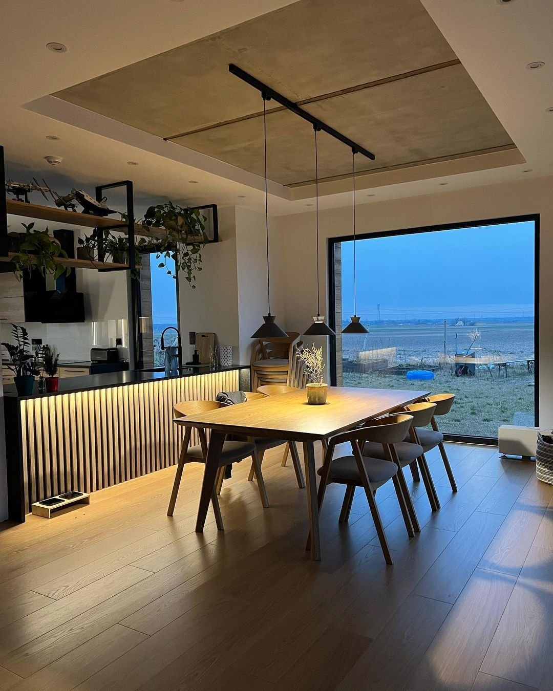A modern dining area with a wooden table and chairs, pendant lights above, and large window offering a view of an outdoor landscape at dusk. Japandi-inspired design ideas bring plants and decor items that create both a functional and beautiful kitchen space.