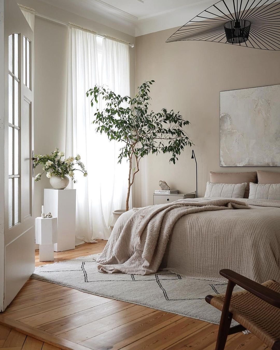 A bright bedroom with a large bed, beige bedding, a tree in a white pot, and minimalist Japandi decor. Sunlight filters through sheer white curtains, illuminating a geometric rug on the wooden floor. This space exemplifies Minimalist Living: How Japandi Decor Can Help Simplify Your Life.