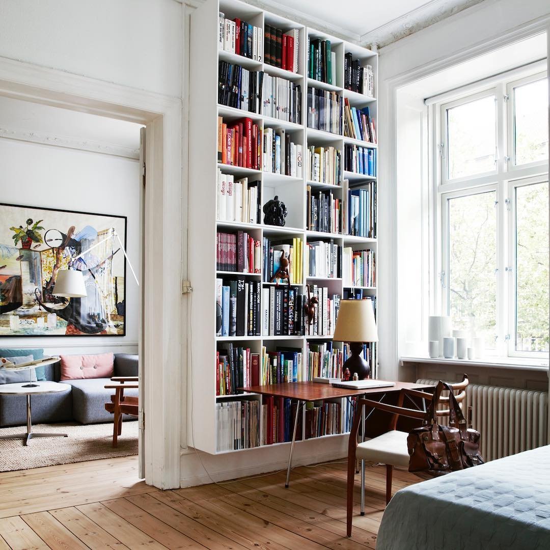 A bright room with a tall, white bookshelf filled with books, a wooden desk with a lamp, and a large window allowing natural light to enter. A sofa is visible in the adjoining room, making it one of the 10 inspiring home office library designs for every decor style.
