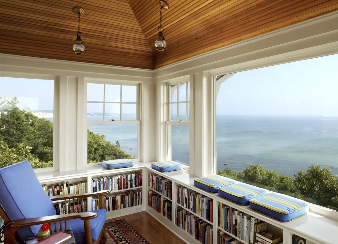 A sunlit room features a wooden ceiling, large windows with ocean views, bookshelves, a blue armchair, and window seats with blue and yellow cushions. This space could easily be one of the 10 Inspiring Home Office Library Designs for Every Decor Style.