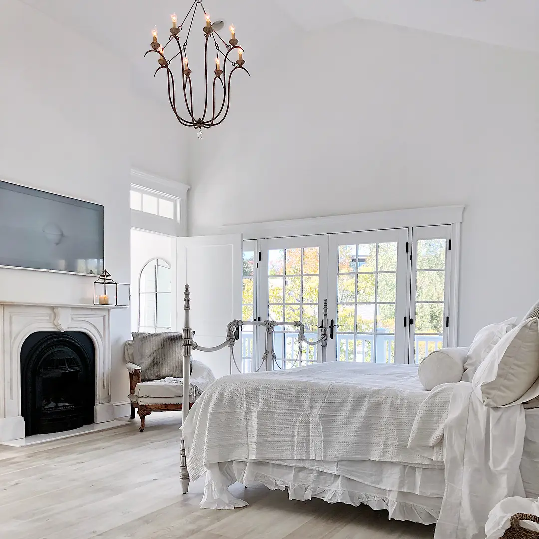 A bright bedroom featuring a white bed, a chandelier, French doors, a fireplace, a wall-mounted TV, and a vintage armchair in a white and beige color scheme perfectly showcases how to embrace rustic elegance to create a French country bedroom retreat.
