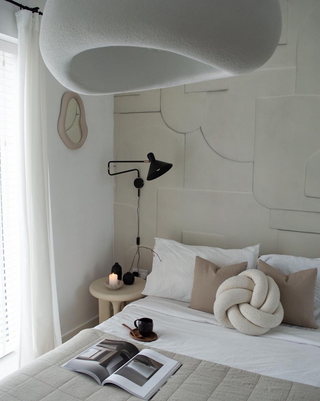 A minimalist bedroom with a white bed, beige pillows, a knotted cushion, a wall-mounted black lamp, a bedside table with a candle and a cup, and an open book. White curtains cover the window. It's an ideal setting for those wanting to know how to incorporate meaningful artifacts in your Japandi bedroom seamlessly.
