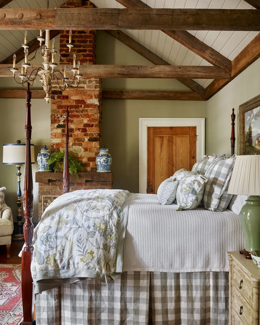 A cozy bedroom with Southern flair features a four-poster bed with plaid bedding, exposed wooden ceiling beams, a brick fireplace, rustic decor, and a chandelier. A wooden door, two blue vases, and a lamp add to the charm in this romantic space.