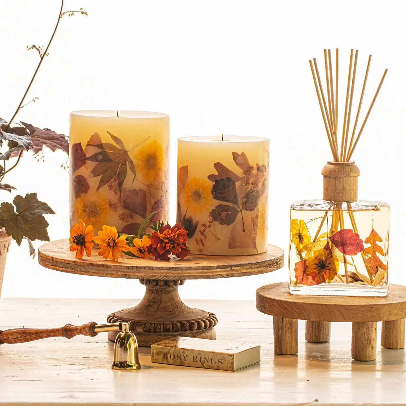 Two candles with embedded dried leaves and flowers sit on a wooden stand beside a reed diffuser and small florals—perfect accents for those exploring how to create a calm bedroom with aromatherapy and fall seasonal scents.