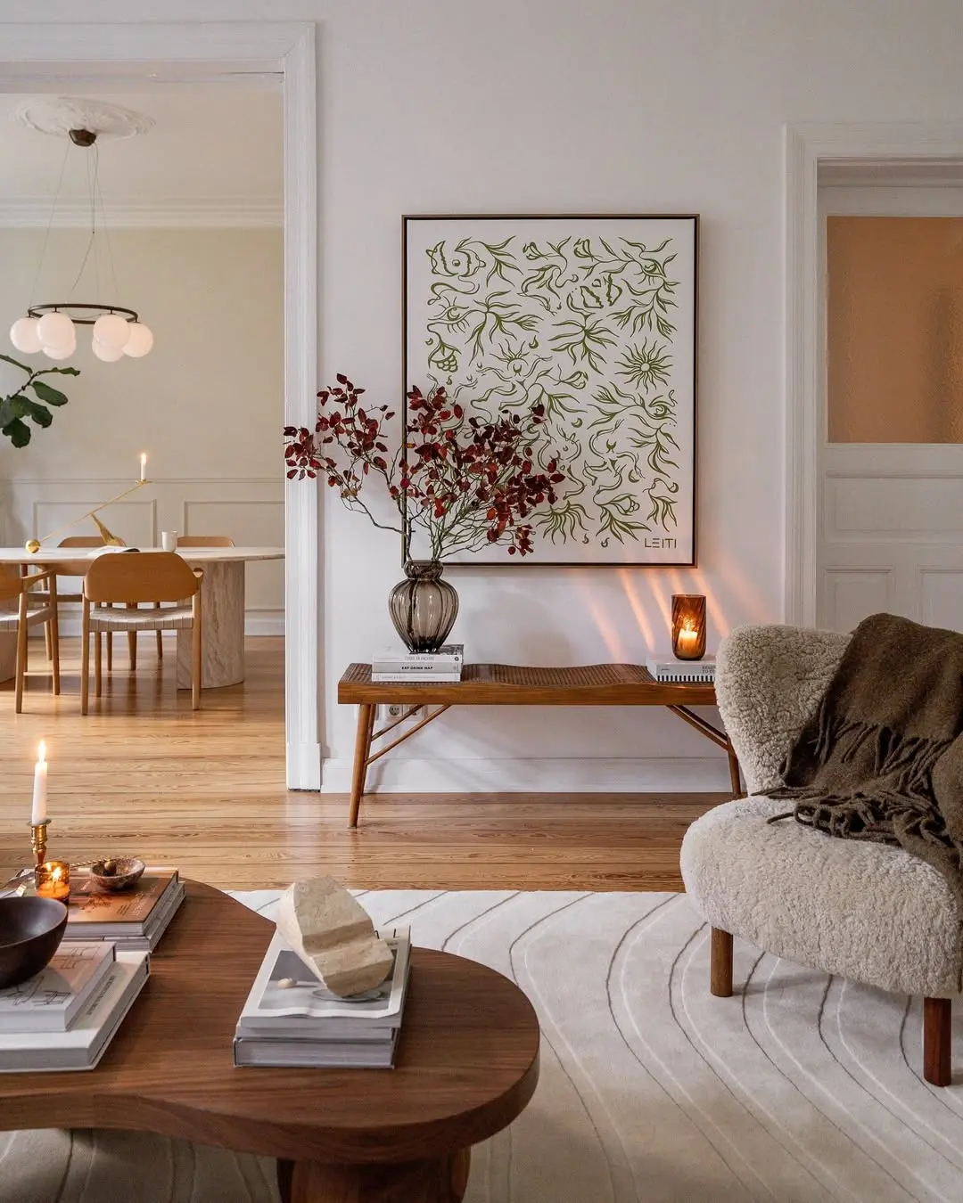 A cozy living room with a round armchair, wooden coffee table, art on the wall, and a bench with a vase of branches shows how to embrace the season: transition your home decor from fall to winter with modern furniture and warm pendant lights.