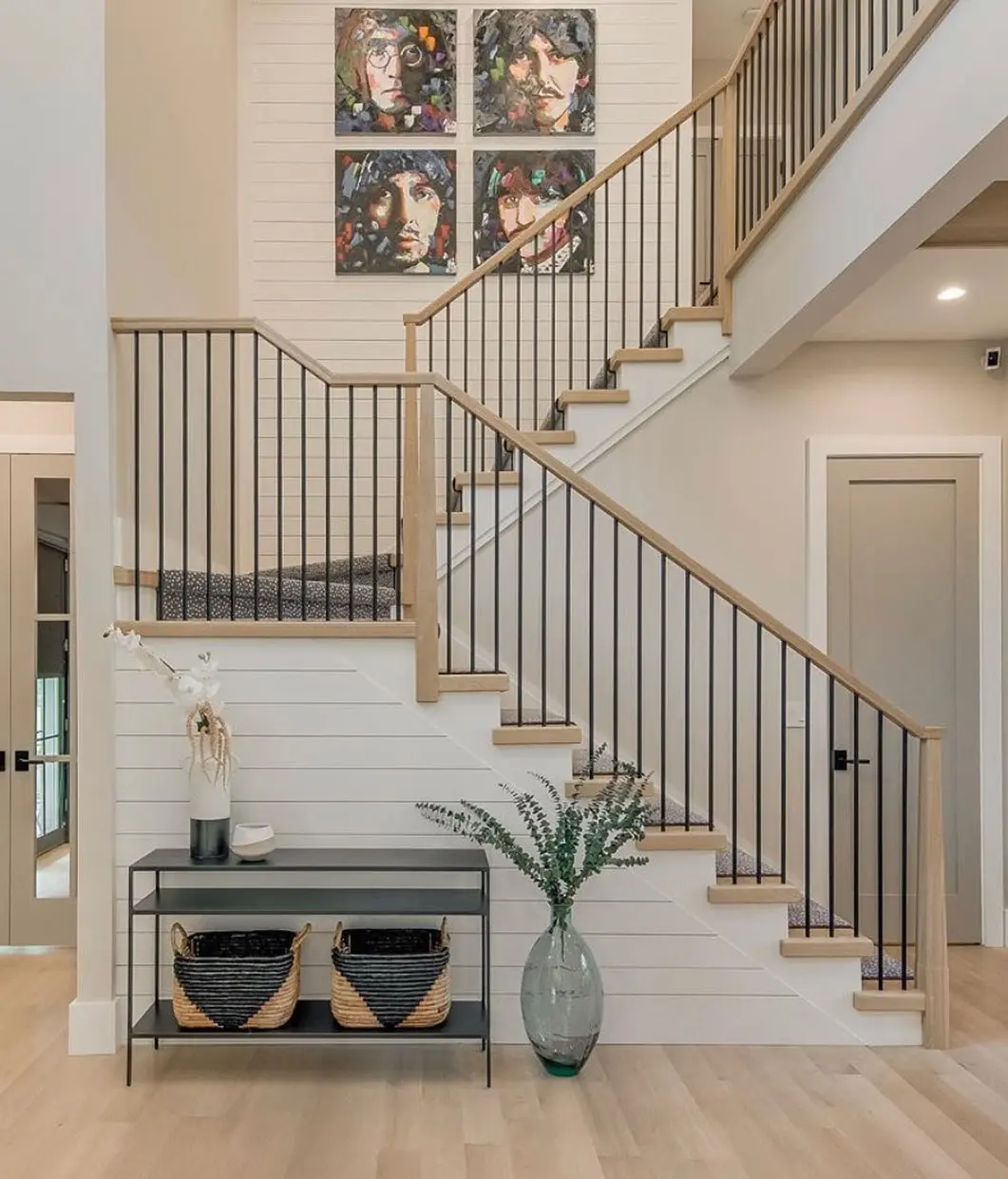 A modern staircase with black railings, light wood accents, and a console table with decor and baskets brings innovative twists to farmhouse stair railing designs. A tall vase with greenery and four colorful portraits complete the look above.