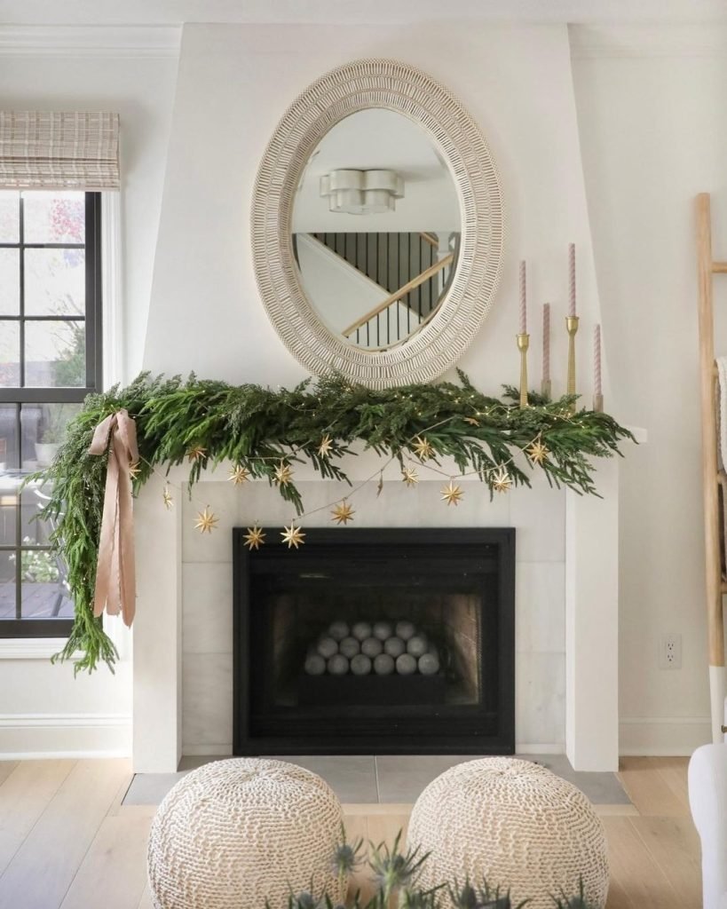 A fireplace decorated with greenery, gold star ornaments, and pink candles stands below a round wicker-framed mirror in a bright living room with light wooden floors and a window with checkered curtains, embracing minimalism: how to create a chic winter decor scheme effortlessly.