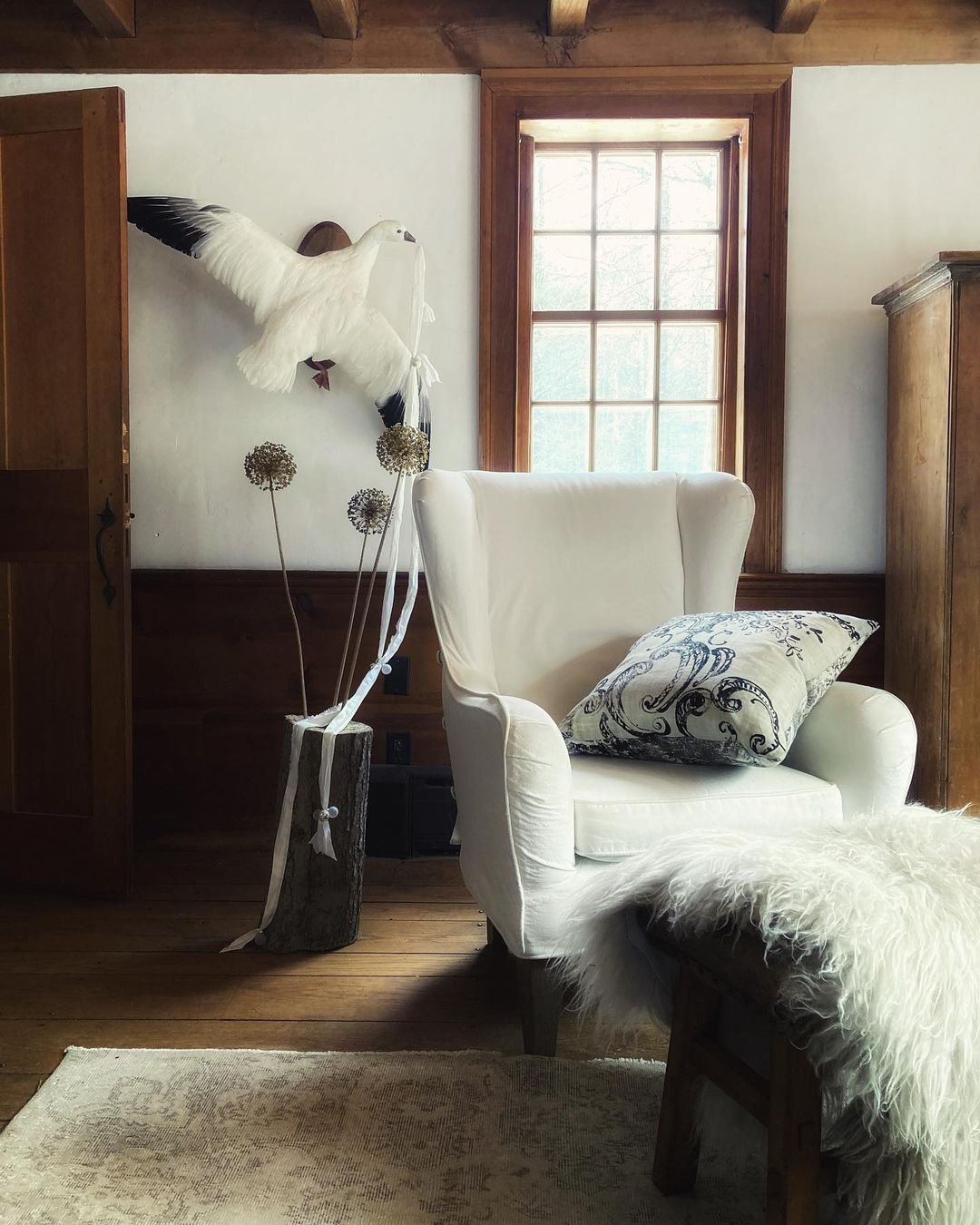A white armchair with a patterned cushion sits in a cozy room with wood accents; above dried flowers in a vase, a taxidermy bird is mounted—showing how slipcover sofas enhance your living room's charm.