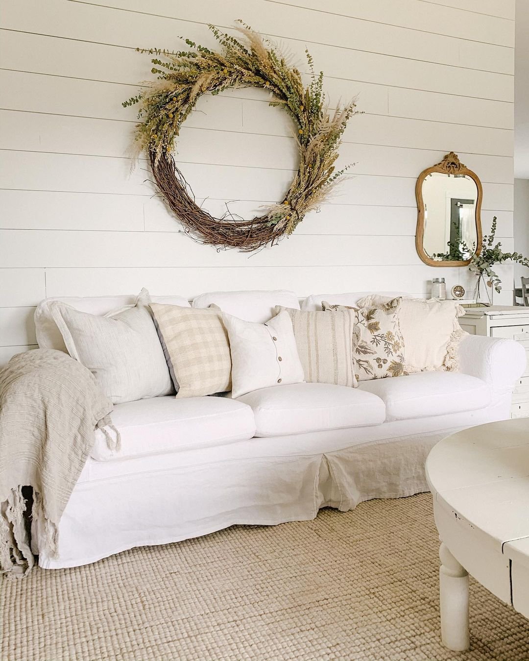 A living room with a white slipcover sofa adorned with neutral-colored pillows and a beige throw blanket. A large rustic wreath hangs on the shiplap wall, and a round mirror and small table are visible nearby. Slipcover sofas effortlessly enhance your living room's charm.
