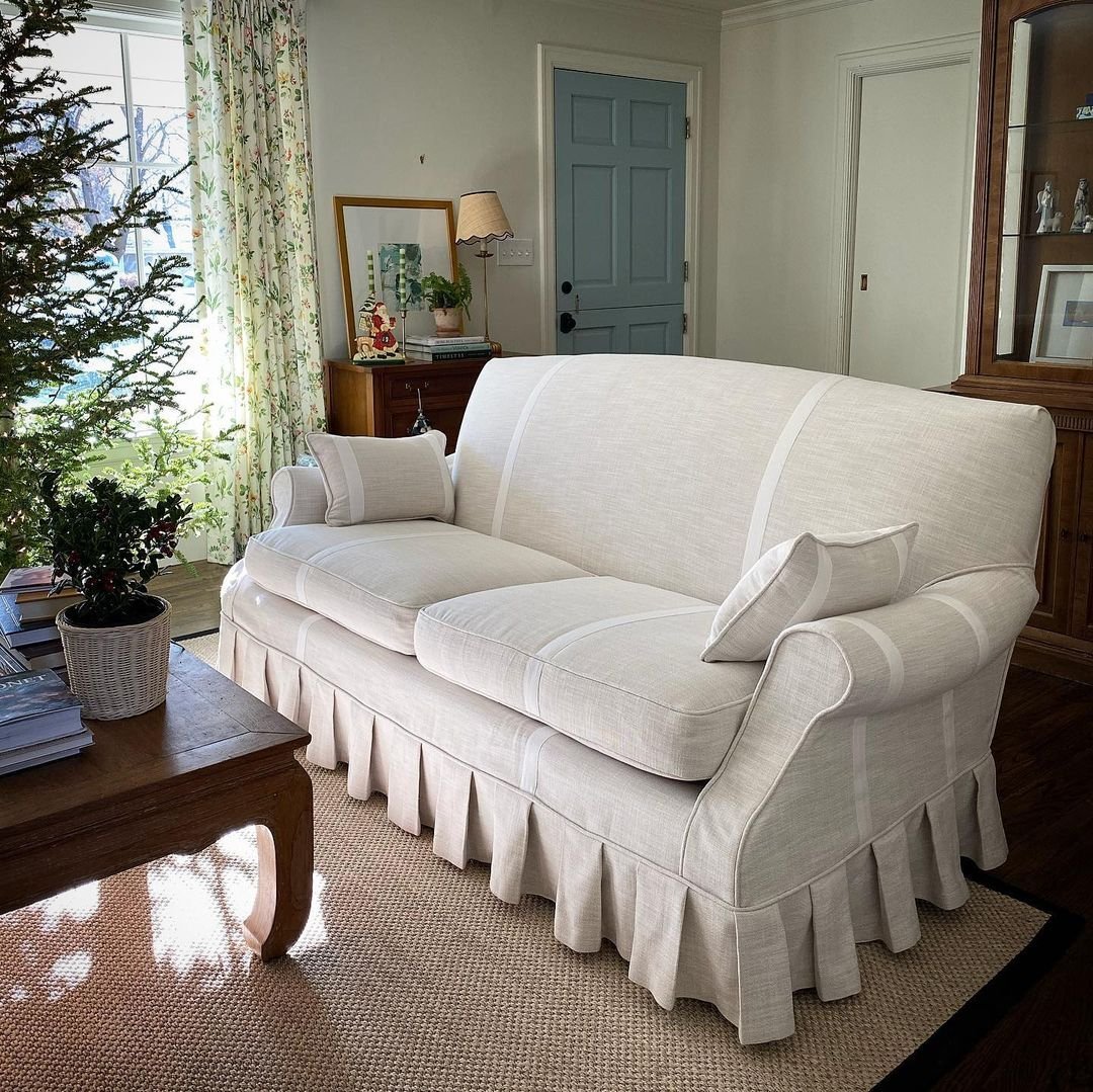 A cozy living room featuring a white slipcover sofa with a ruffled skirt, wooden coffee table with plants and books, a blue door, and a window with floral curtains. A Christmas tree stands in the left corner, showing how slipcover sofas enhance your living room's charm.