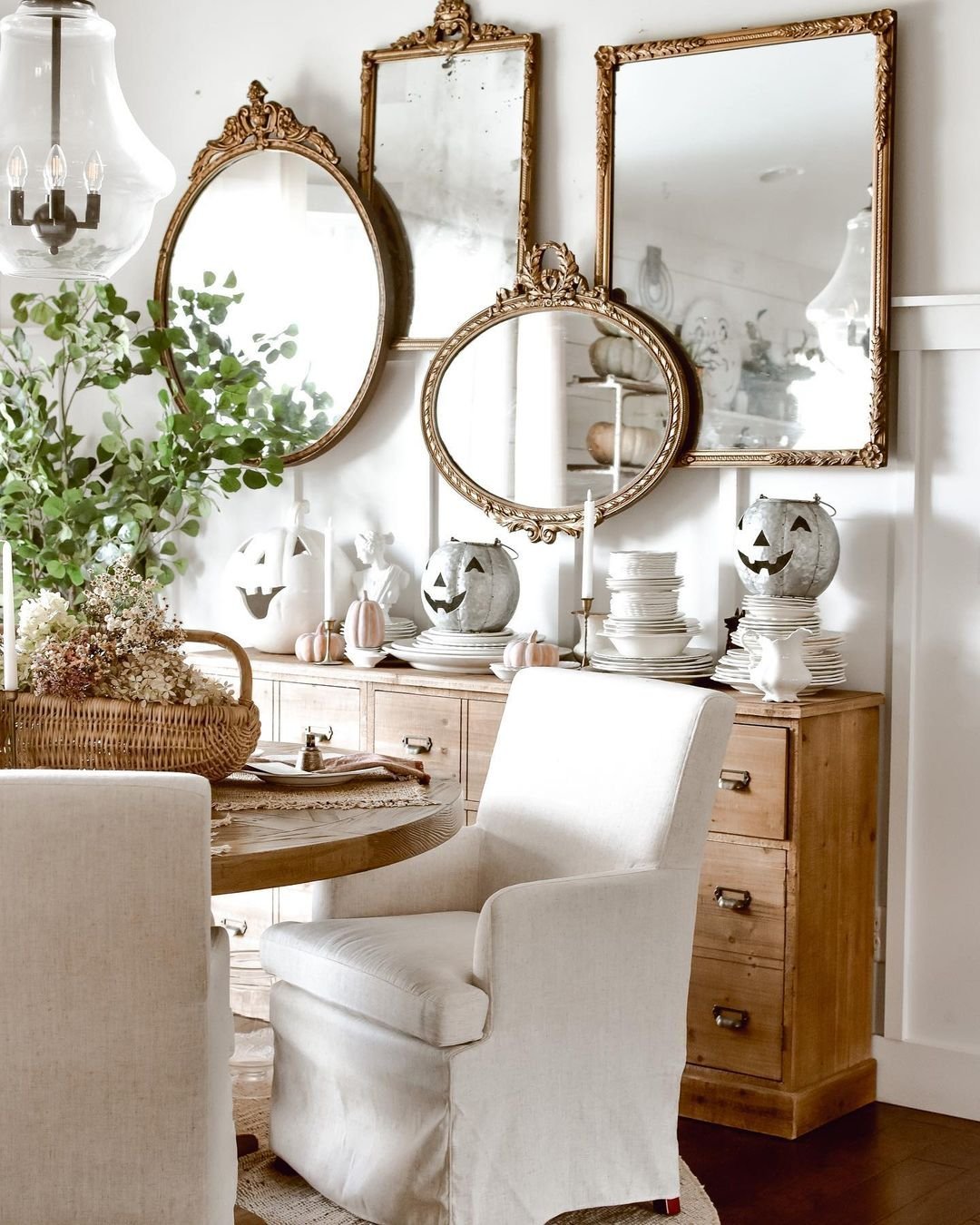 A dining room features a round table, white chairs, a wooden sideboard, and Halloween decorations including pumpkin lanterns. The room is decorated with greenery and ornate mirrors on the wall. Consider adding slipcover sofas for versatility and added charm to complement this festive space.