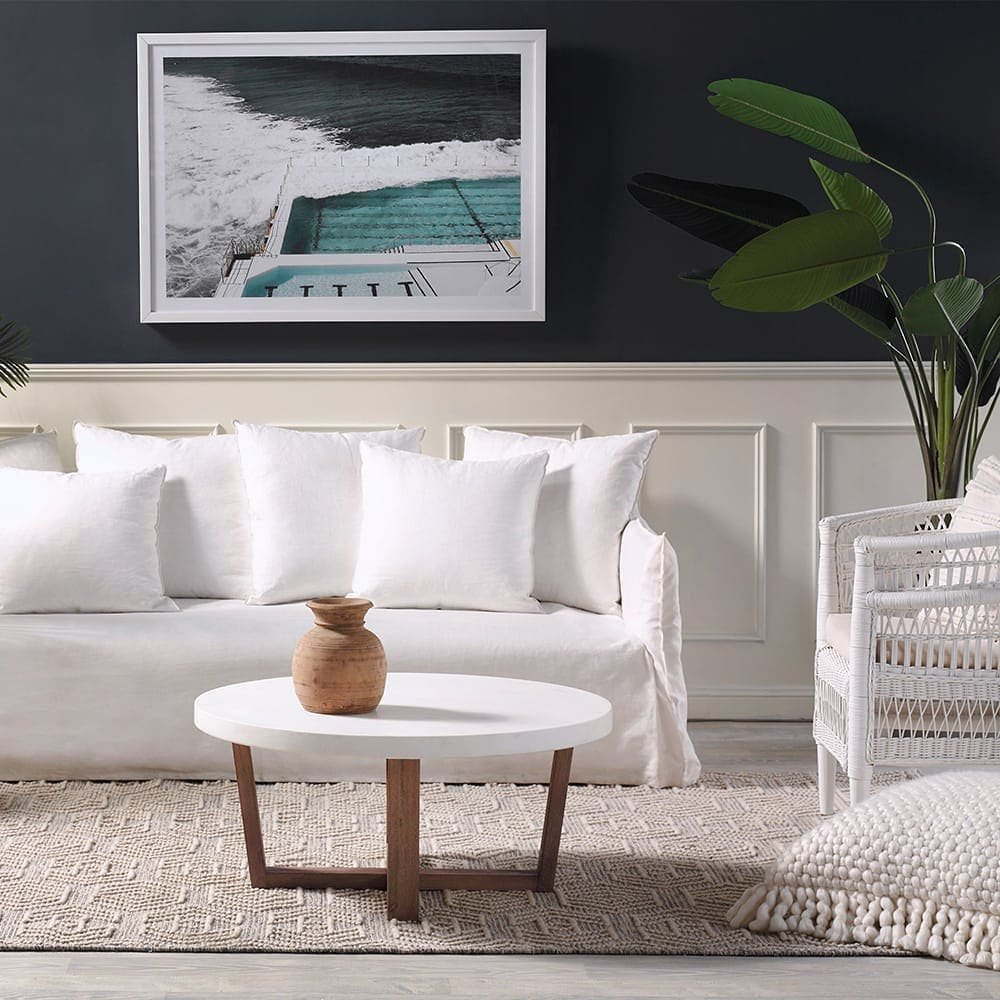 A living room with a white slipcover sofa, wicker chair, round coffee table with a vase, and framed ocean-themed artwork. The wall is dark with paneled wainscoting, and a large green plant is on the right. How Slipcover Sofas Enhance Your Living Room's Charm is clearly demonstrated in this cozy space.