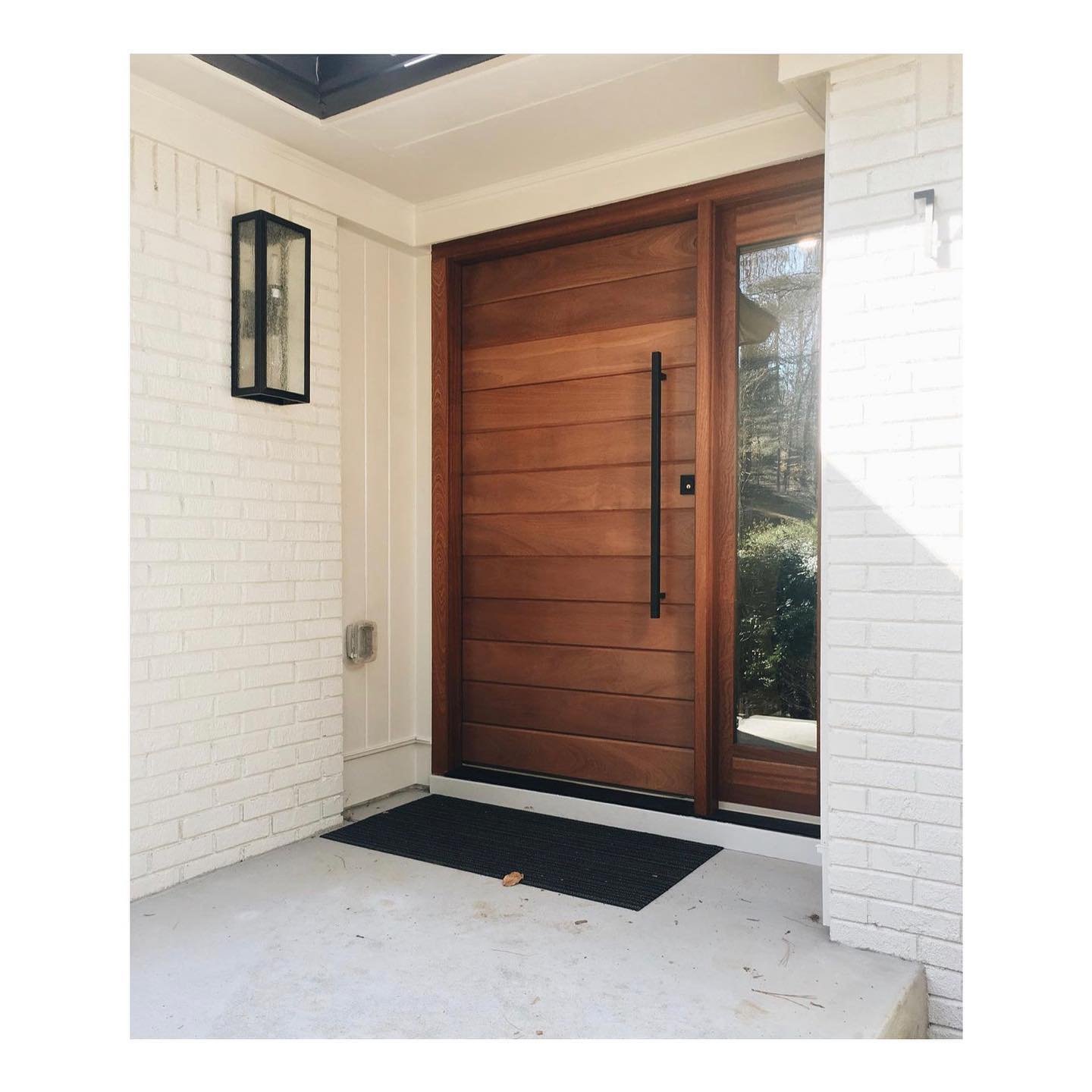 A solid wood front door with a vertical glass panel, flanked by white painted brick walls and a black outdoor wall light, leads to a small concrete porch with a black doormat, showcasing the perfect blend from classic to contemporary.